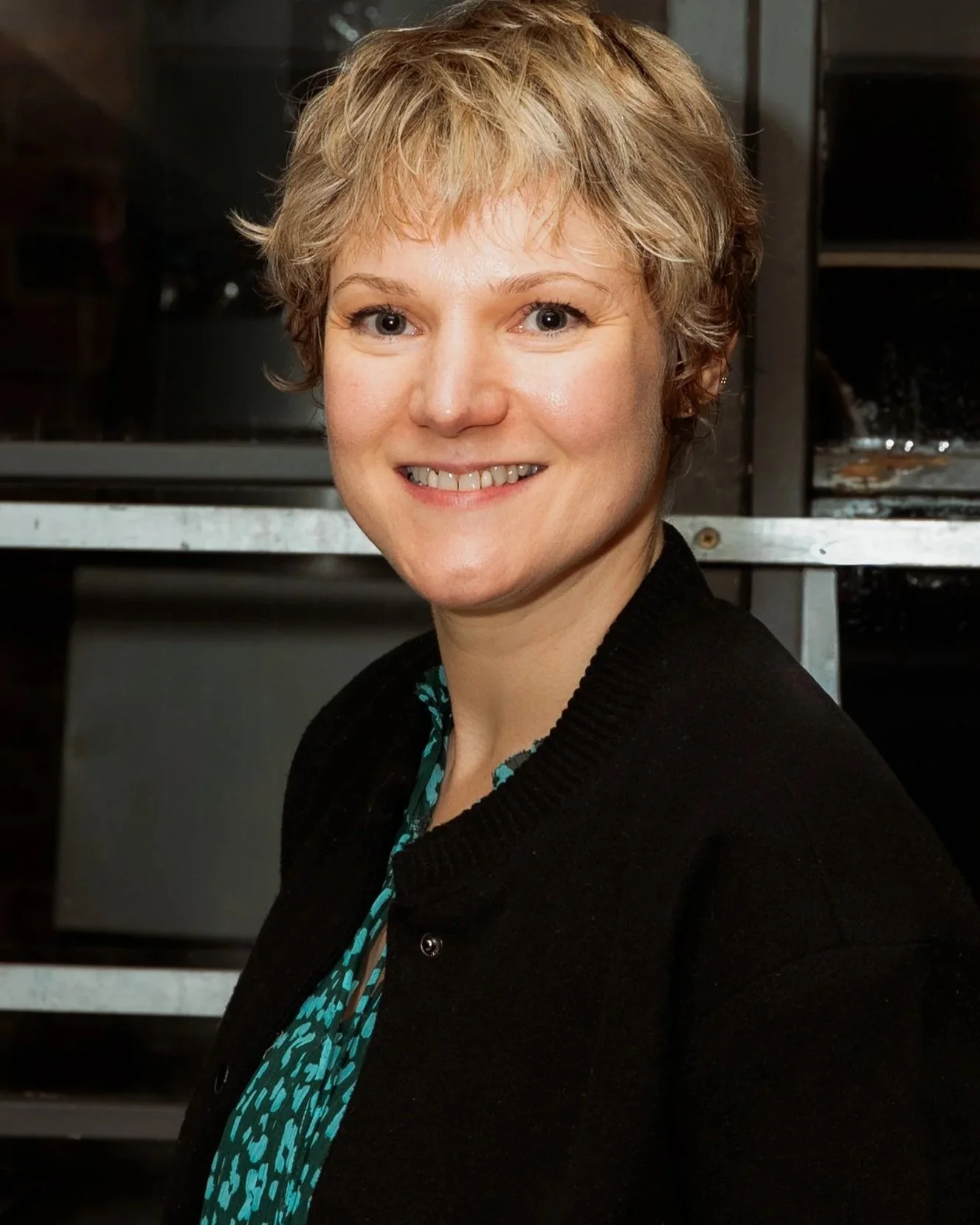 Portrait of a woman with short blonde hair wearing a black jacket and teal dress, smiling in an indoor setting with black and metallic furniture in the background.