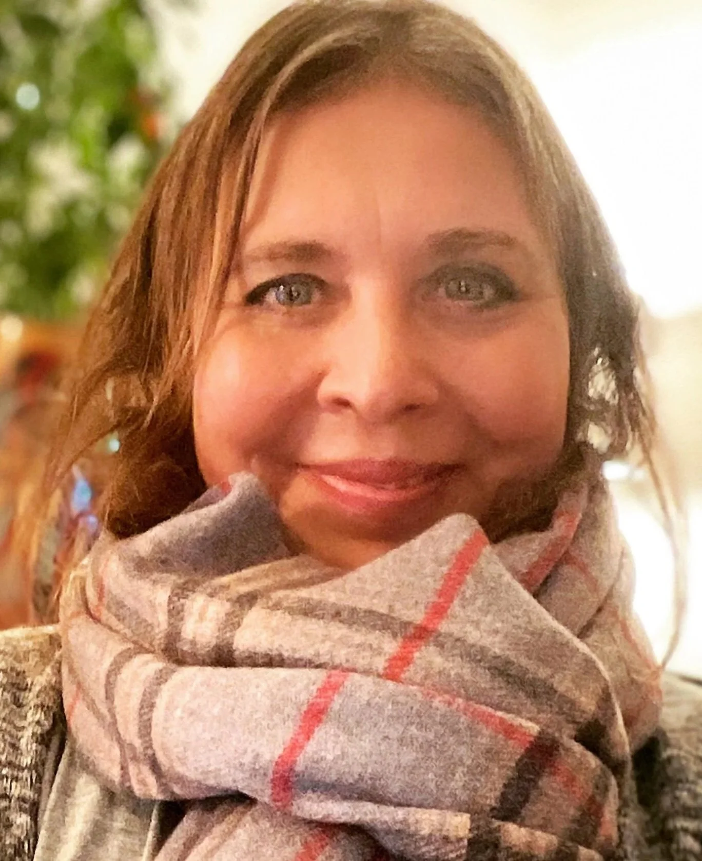A woman with light brown hair, wearing a plaid scarf, smiling in a warmly-lit indoor setting with a blurred background.