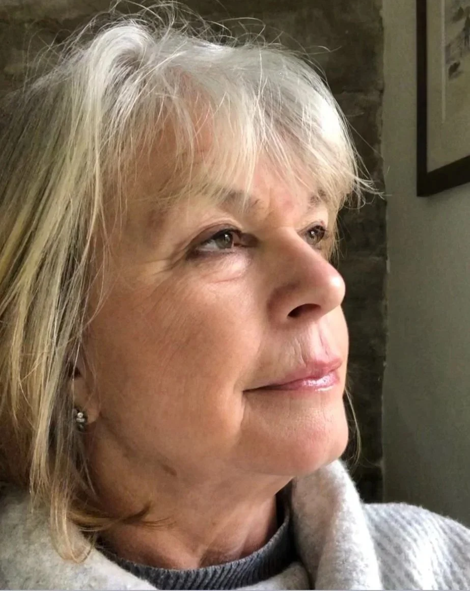 Close-up of an older woman with blonde hair, wearing a light-colored sweater, looking thoughtfully into the distance indoors.