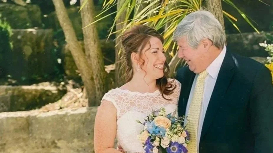 A bride and an older man, possibly her father, smiling and sharing a joyful moment outside during a wedding ceremony. The bride is holding a bouquet of flowers and wearing a lace wedding dress. The background features trees and greenery.