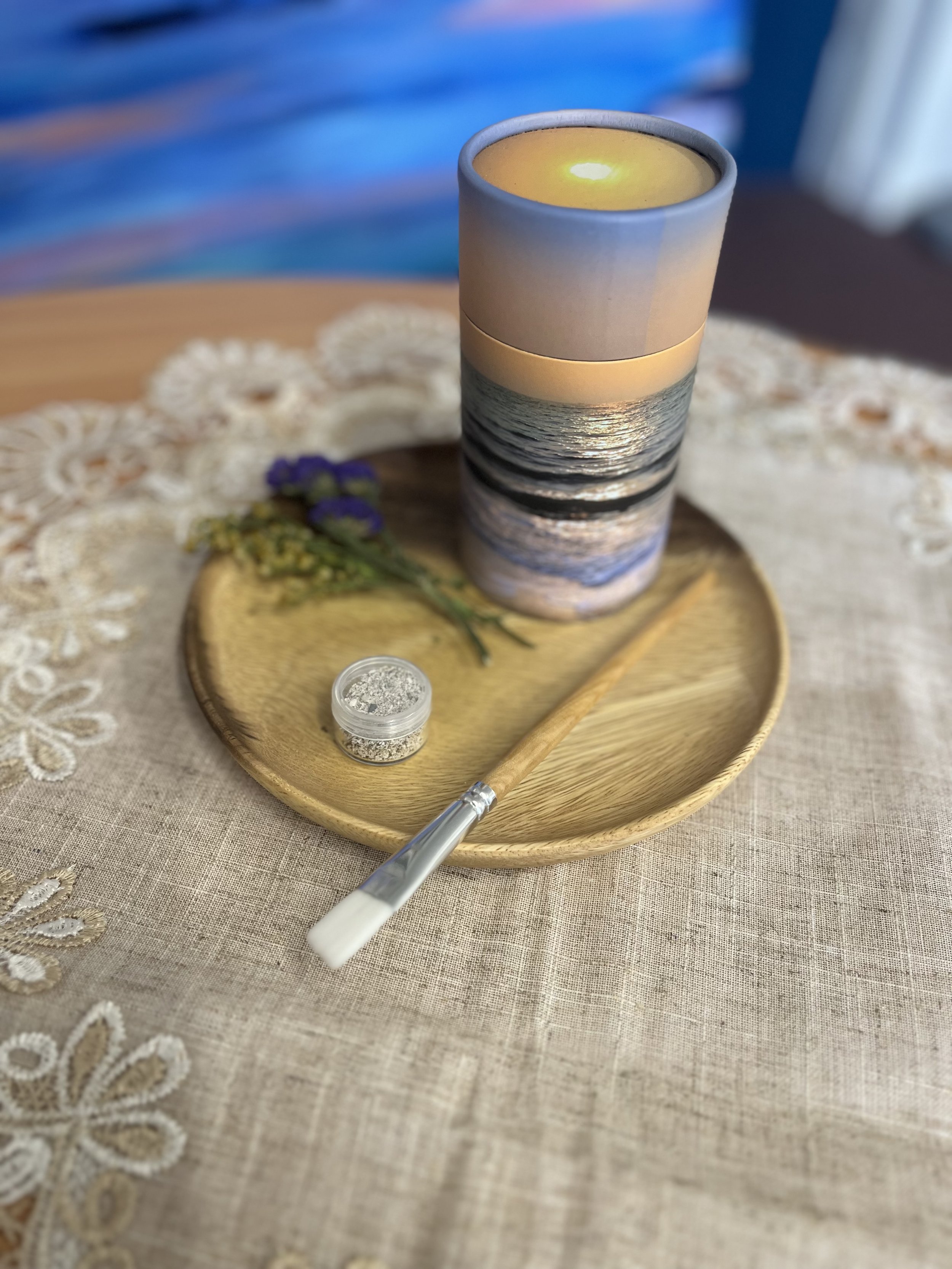 A lit candle in a gray ceramic holder, placed on a wooden tray with lavender sprigs and a small container of silver glitter, all on a beige embroidered tablecloth.