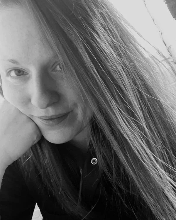 A young woman with long, straight hair resting her face on her hand, looking at the camera with a slight smile, in black and white.
