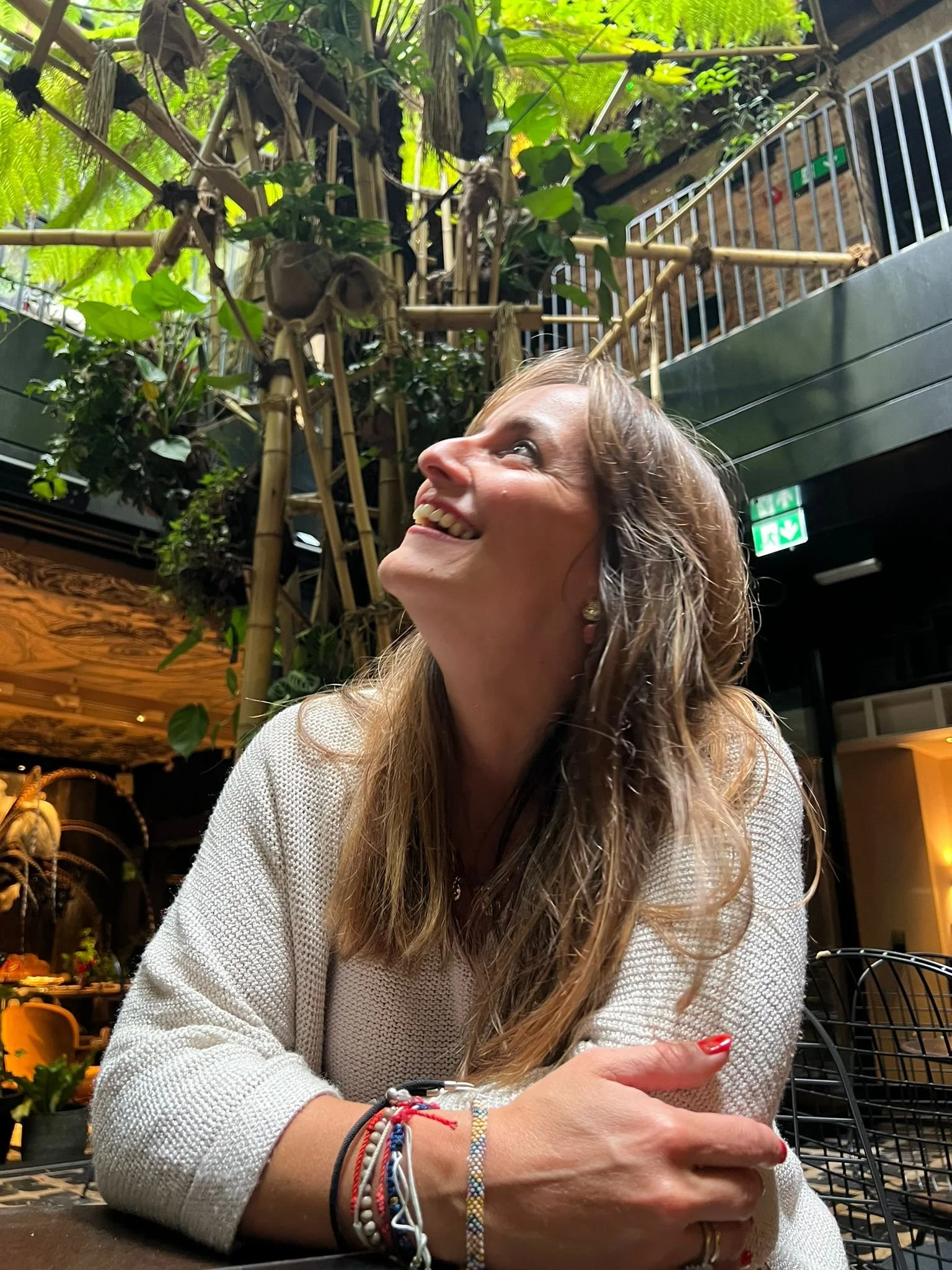 A smiling woman with long, curly hair sitting at a table inside a lush, green environment. She has multiple colorful bracelets on her wrist, red nail polishand is looking up with a joyful expression. The background features vibrant plants and bamboo decor.
