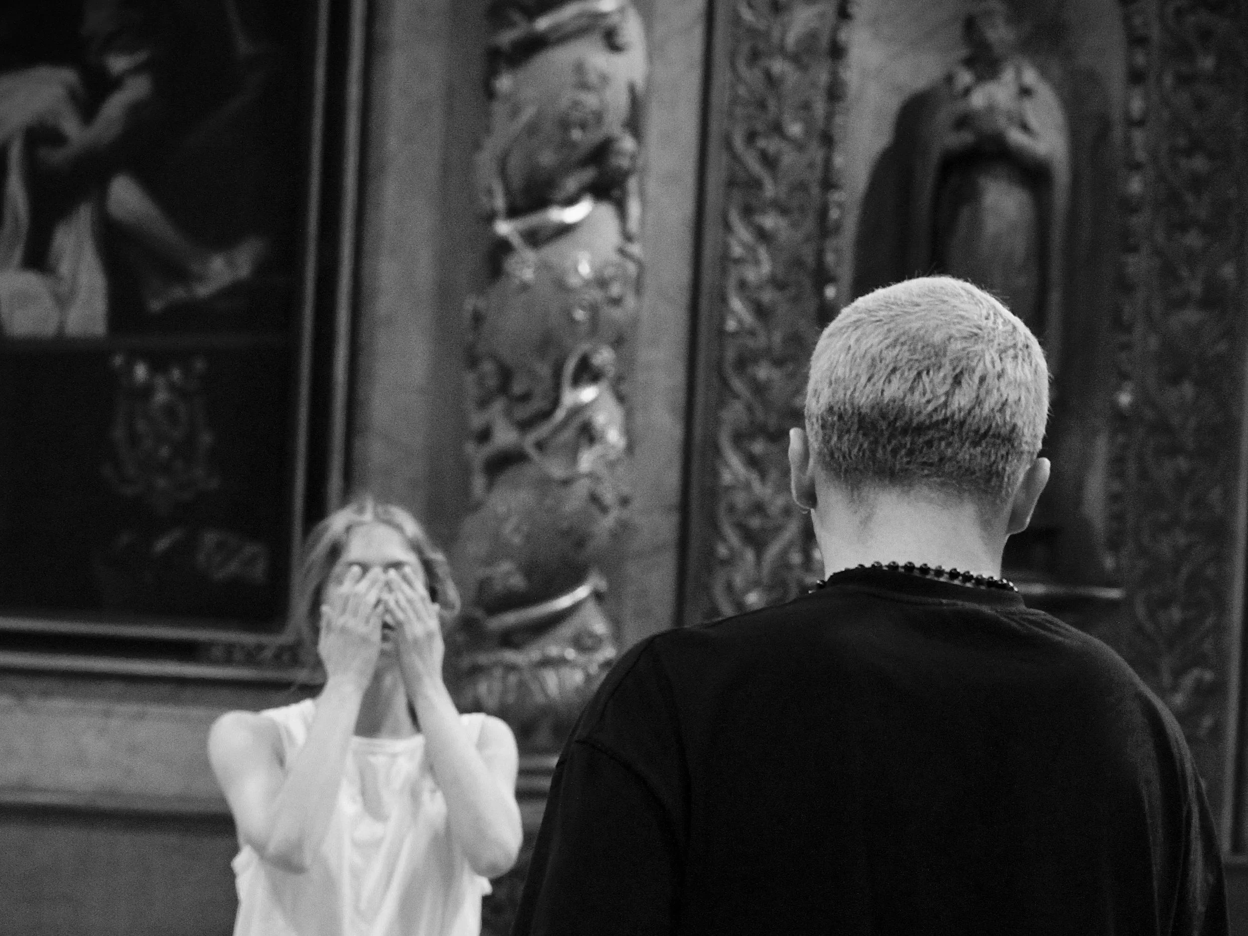 Un garçon avec des cheveux rasés de couleur blonde ou grise interagit avec une sculpture d'une femme en céramique ou en porcelaine, dans une pièce avec des décorations murales. La photo est en noir et blanc.