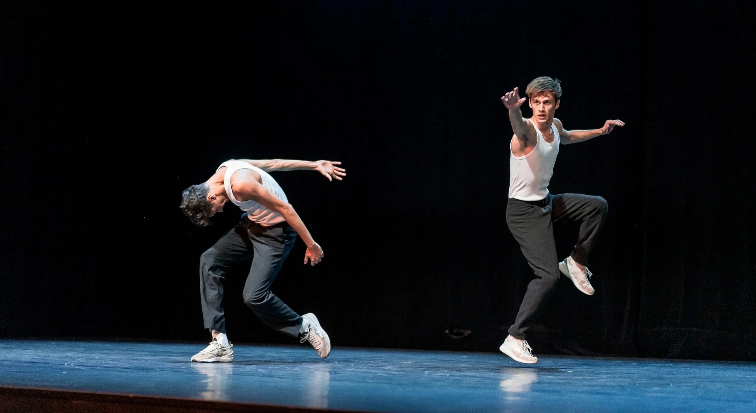 Deux danseurs masculins en mouvement sur scène, portant des vêtements décontractés, dans une performance de danse contemporaine, avec un fond noir.