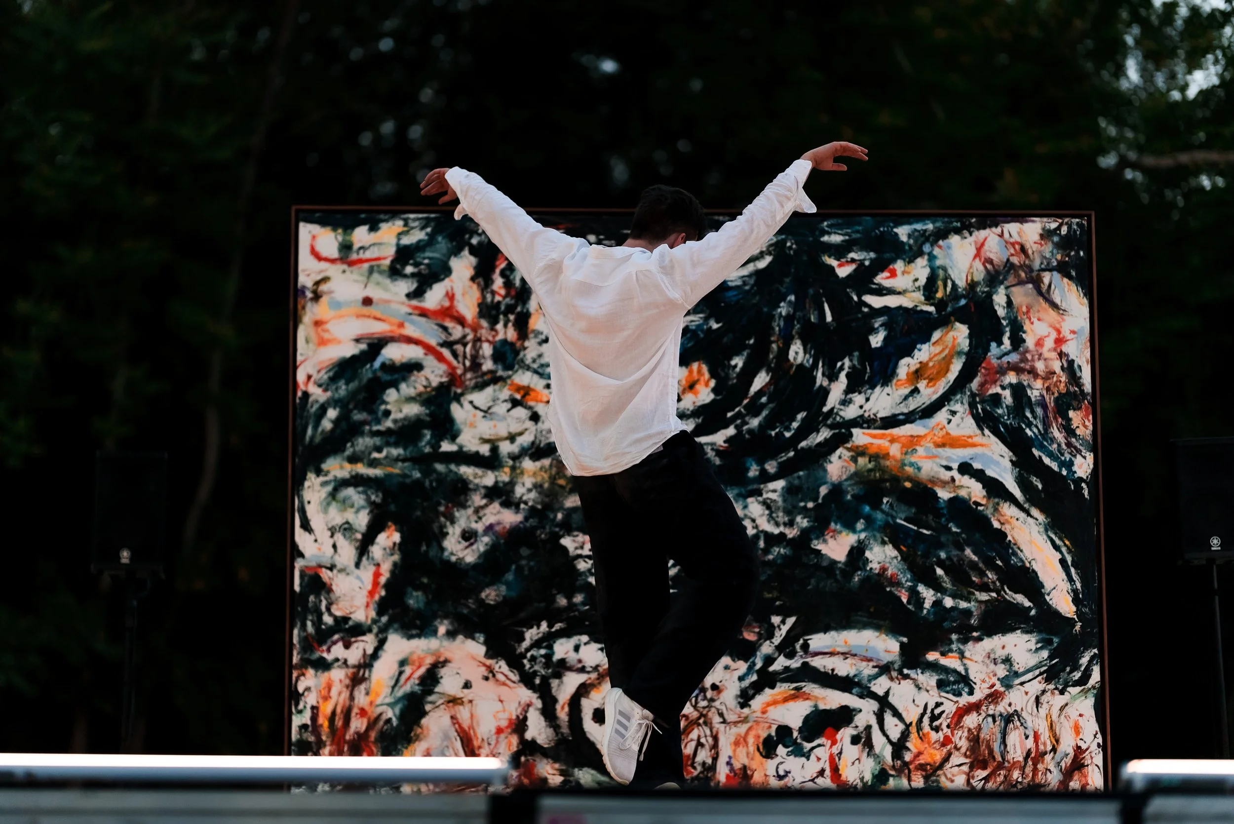 Un jeune homme danse devant une peinture abstraite lors d'un événement en plein air. Il porte un sweat blanc, des pantalons noirs et des baskets blanches. La scène est entourée d'arbres et l'atmosphère est artistique et dynamique.