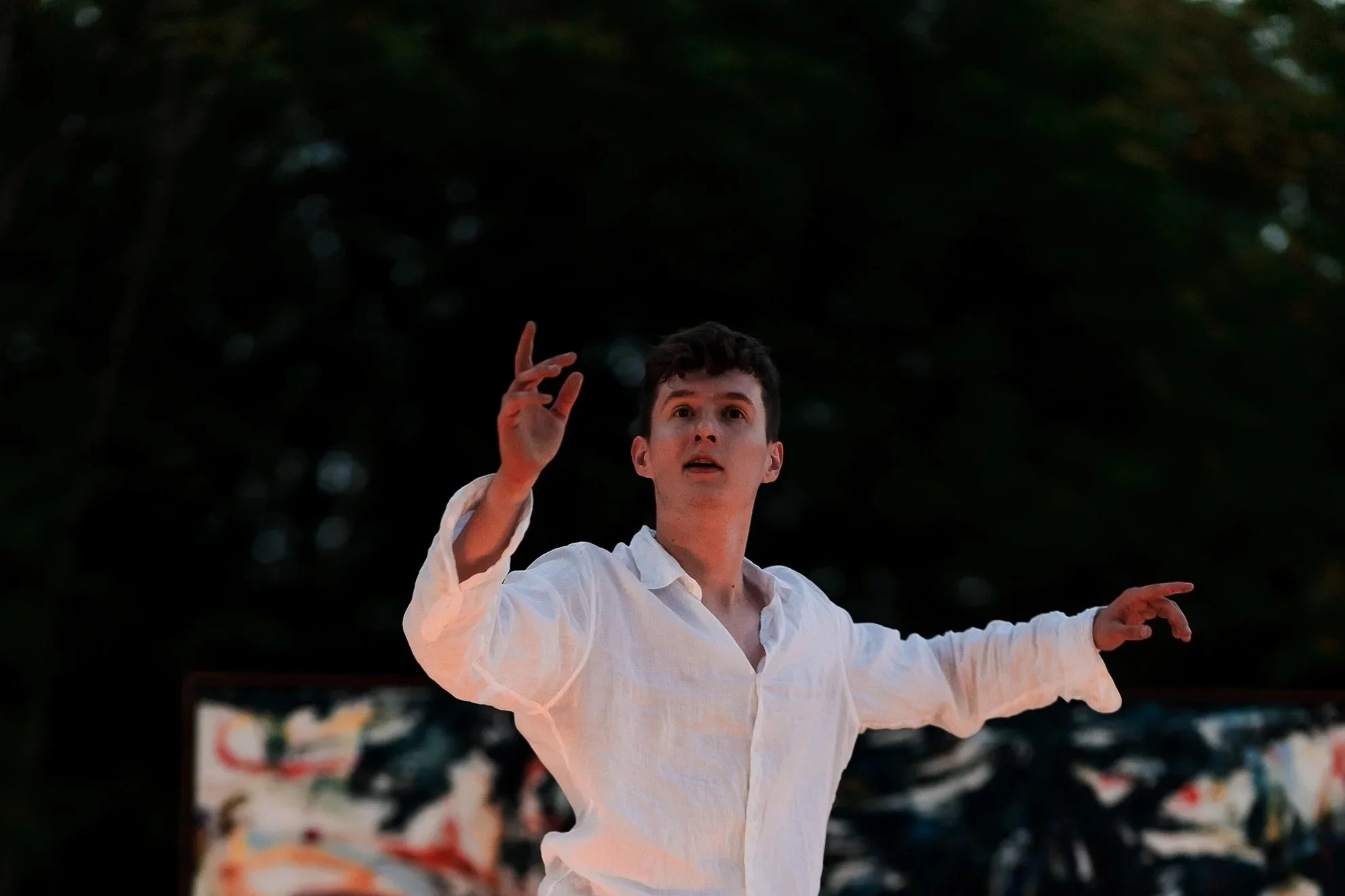 Jeune homme en chemise blanche, gestes gracieux, fond flou d'un arbre et d'une scène de performance 