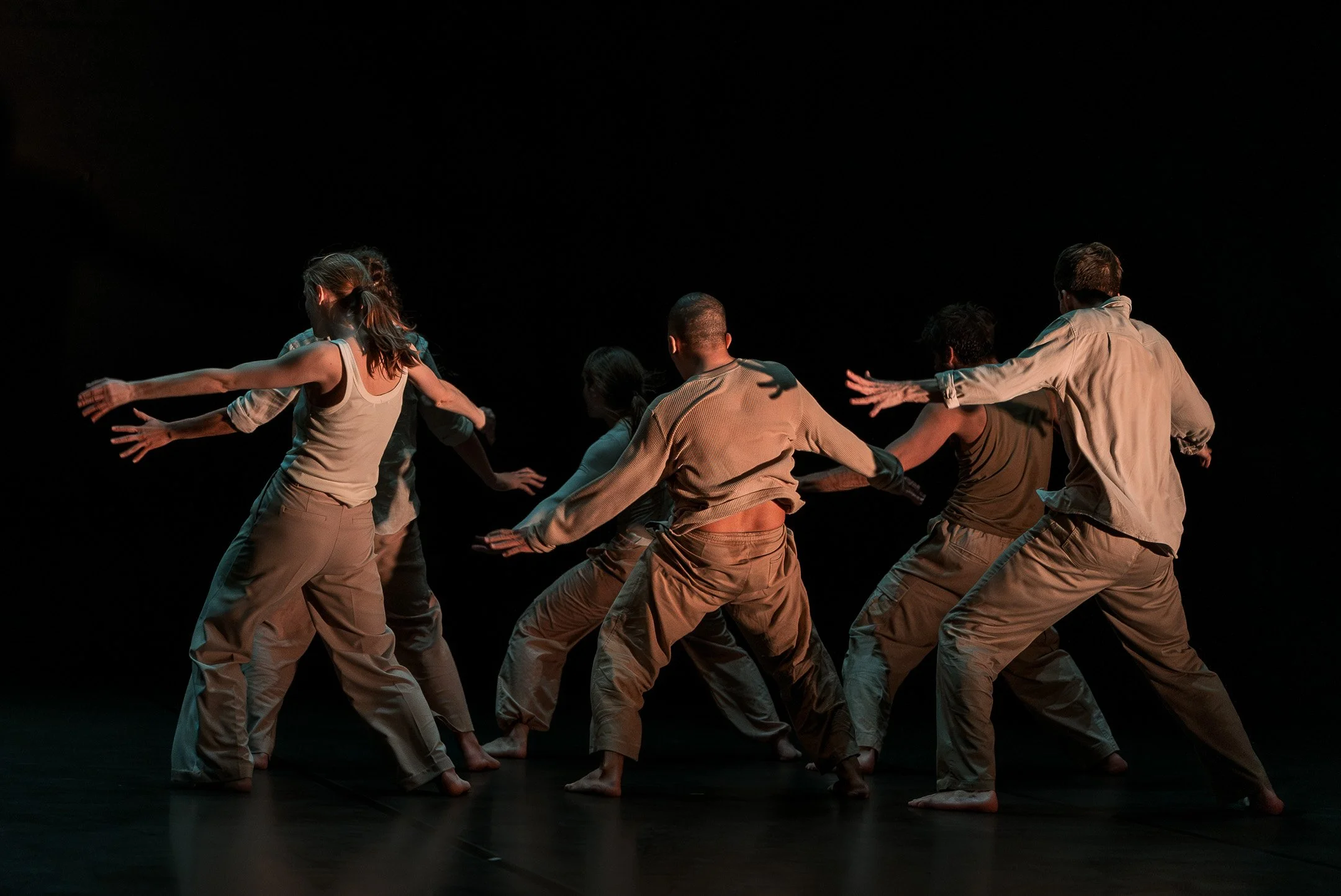 Groupe de danseurs en costume beige, en mouvement dans une performance contemporaine, dans une scène sombre.