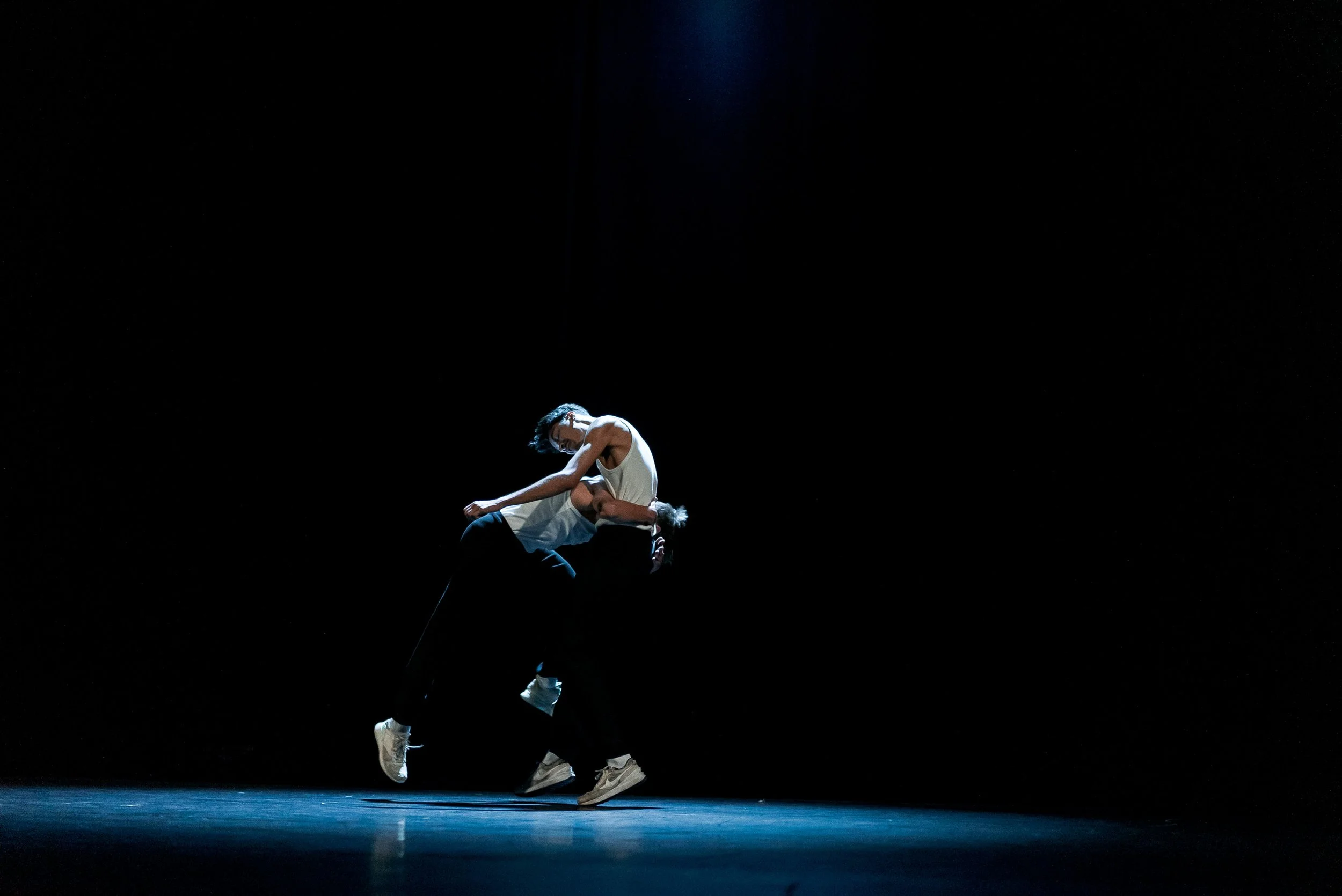 Deux danseurs modernes en tenue décontractée effectuent une performance acrobatique sur scène sombre, avec un éclairage concentré sur eux.