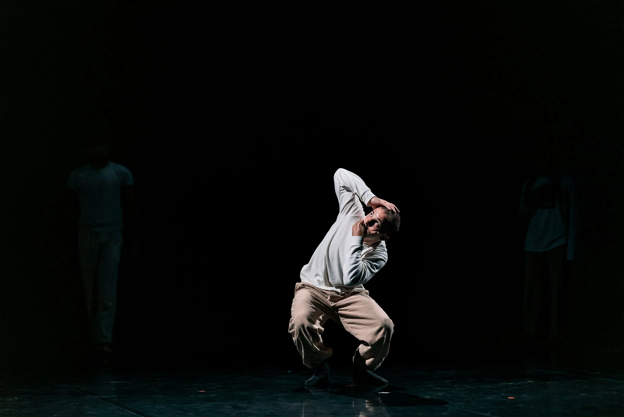 Un danseur en tenue décontractée effectue une pose sur scène sombre, avec deux autres personnes visibles en arrière-plan.