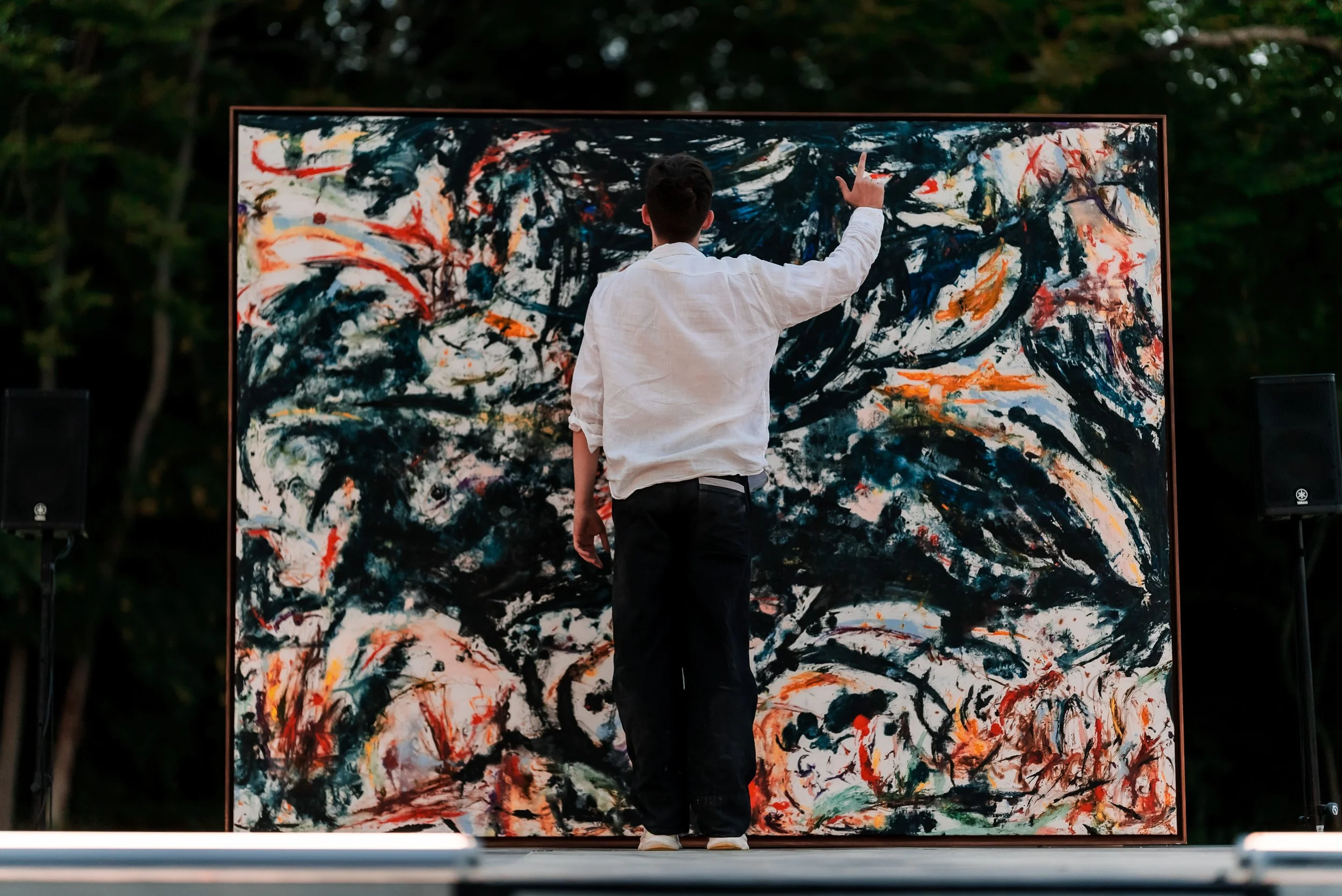 Un homme regarde une grande toile abstraite colorée devant un public, en extérieur, lors d'un événement artistique.