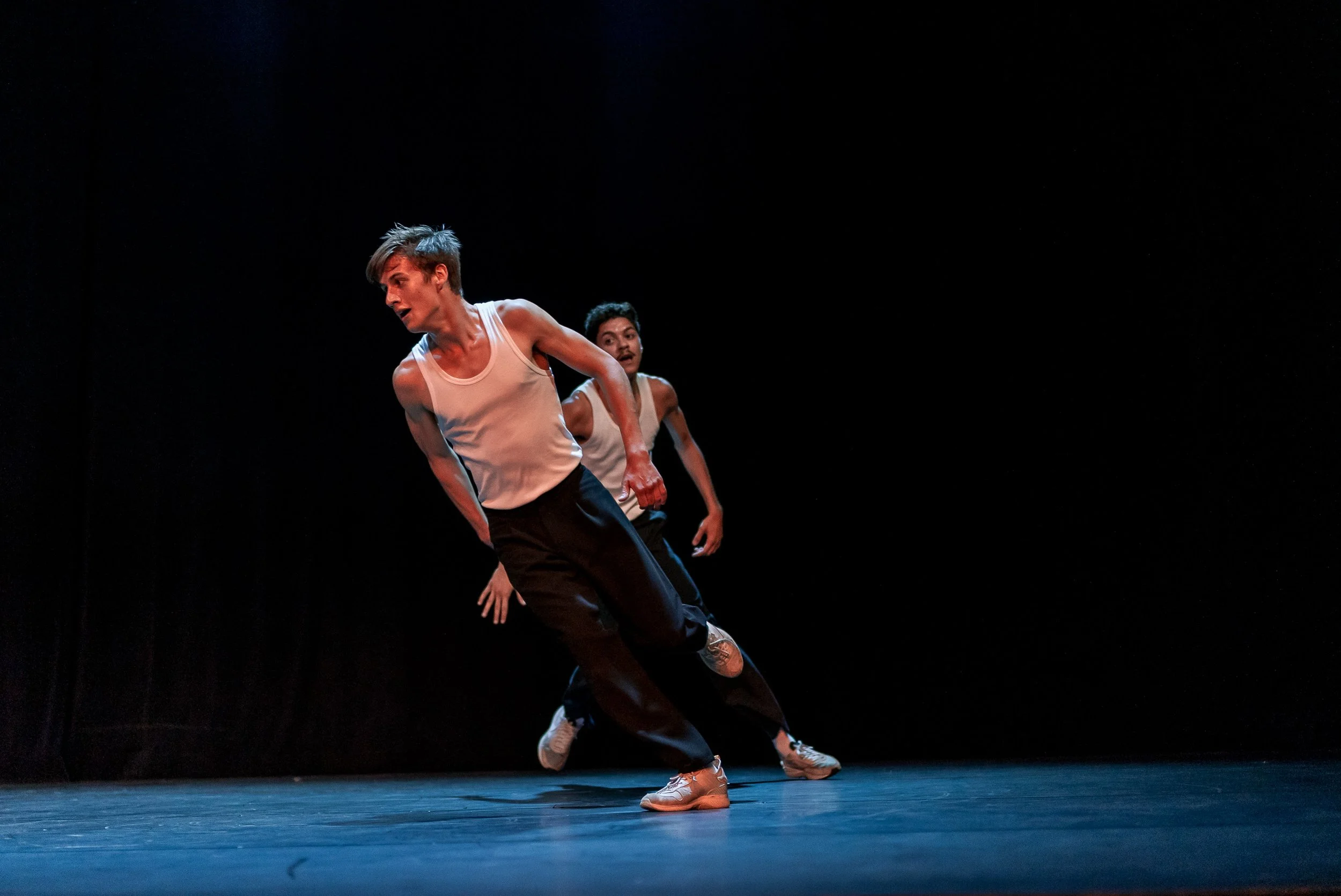 Deux danseurs masculins en mouvements sur scène, portant des débardeurs blancs et des pantalons noirs, avec un fond noir minimaliste.