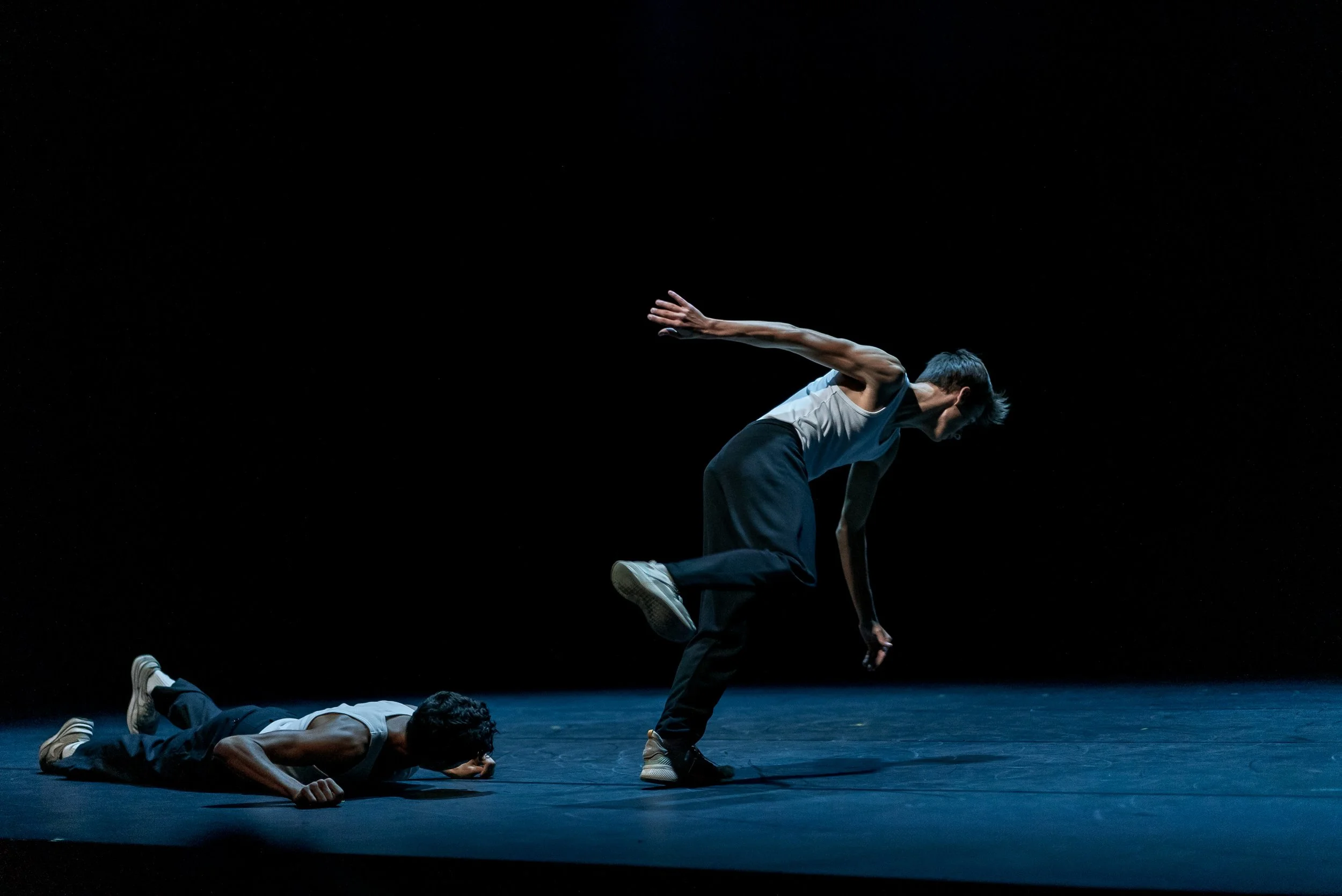 Deux danseurs exécutent une performance de danse contemporaine sur scène sombre, deux allongés au sol et un en position de plié, tous portant des vêtements décontractés. Le fond est noir, mettant en valeur leurs mouvements.