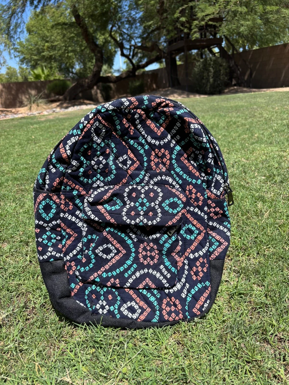 Colorful patterned backpack on grass with trees and a wooden structure in background.