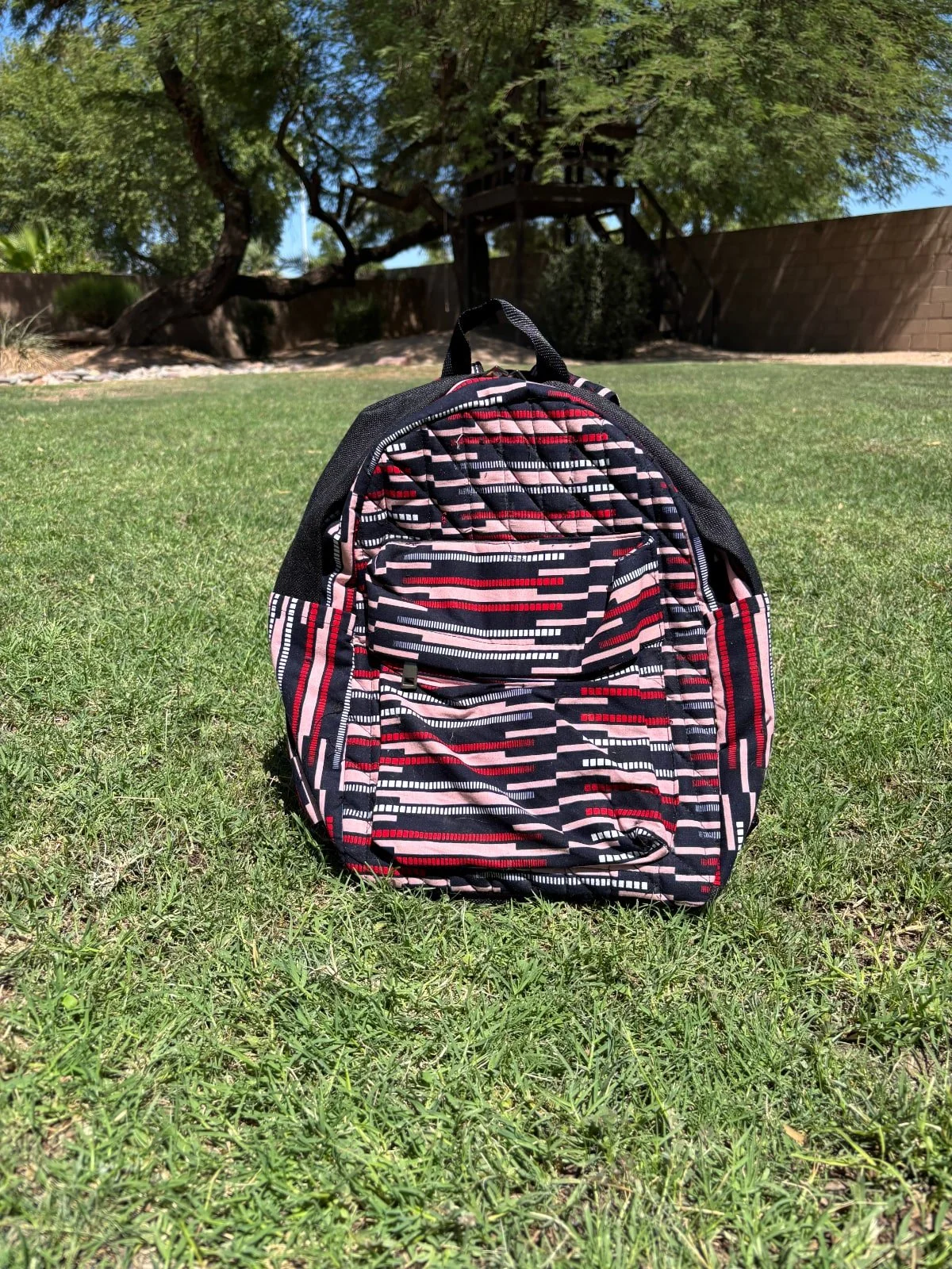 A backpack with a geometric black, red, pink, and white pattern resting on green grass in a park with trees and a wooden structure in the background.