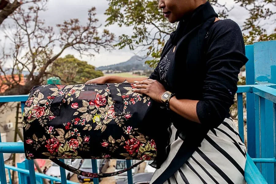 A woman standing on a blue railing outdoors, holding a floral-patterned bag with a cityscape and mountains in the background.