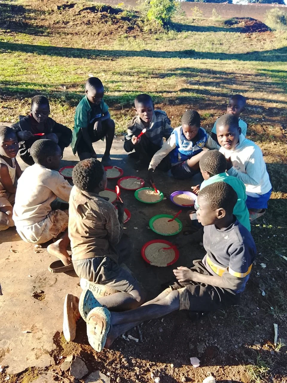 Group of children sitting on the ground outdoors, playing with round plates and red spoons.