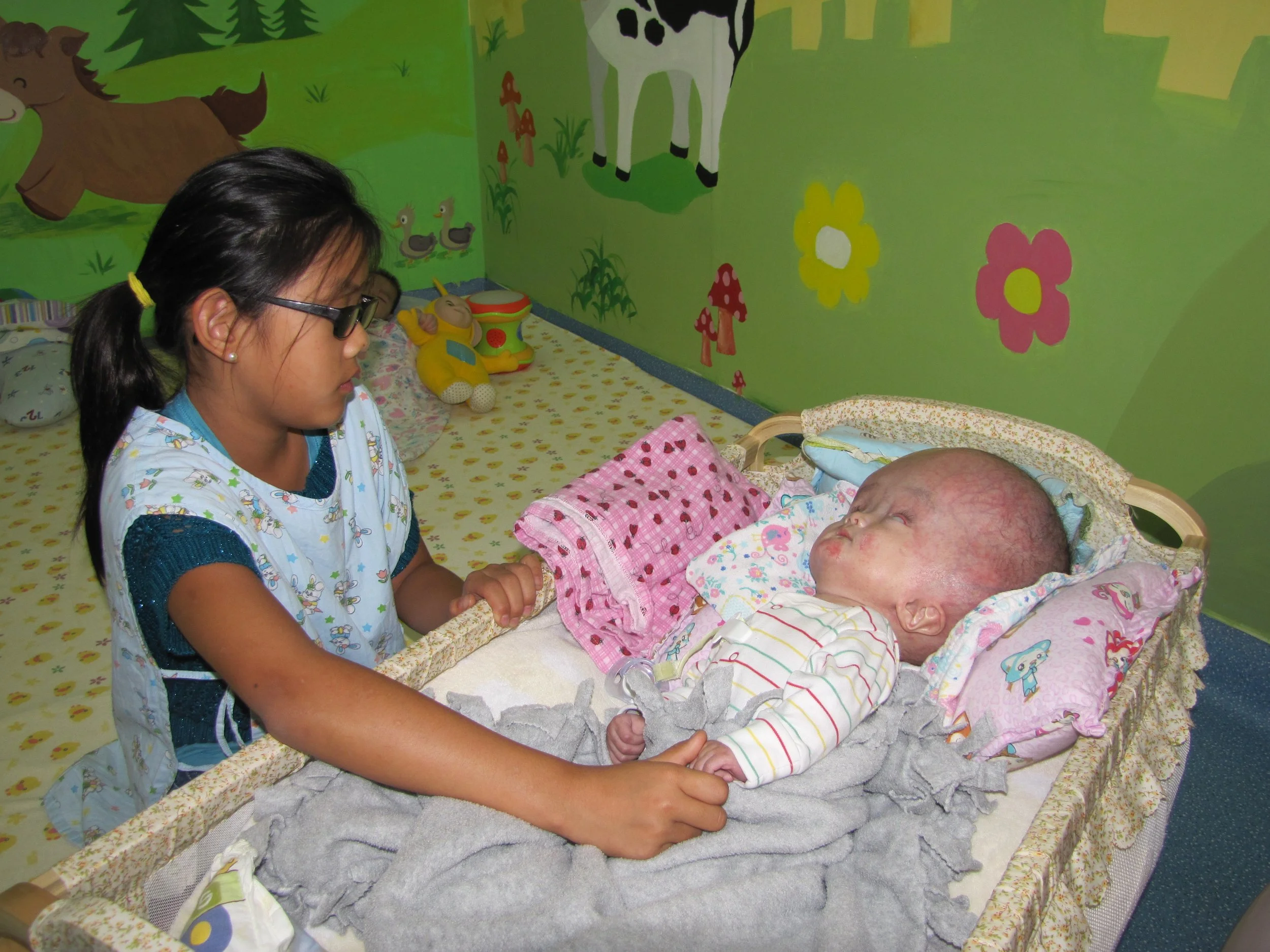 A young girl with glasses, black hair tied in a ponytail with a yellow hair tie, sitting beside a baby in a crib. They are holding hands. The background features a brightly painted green wall with cartoon animals, flowers, and mushrooms. The crib has patterned bedding with a pink blanket.