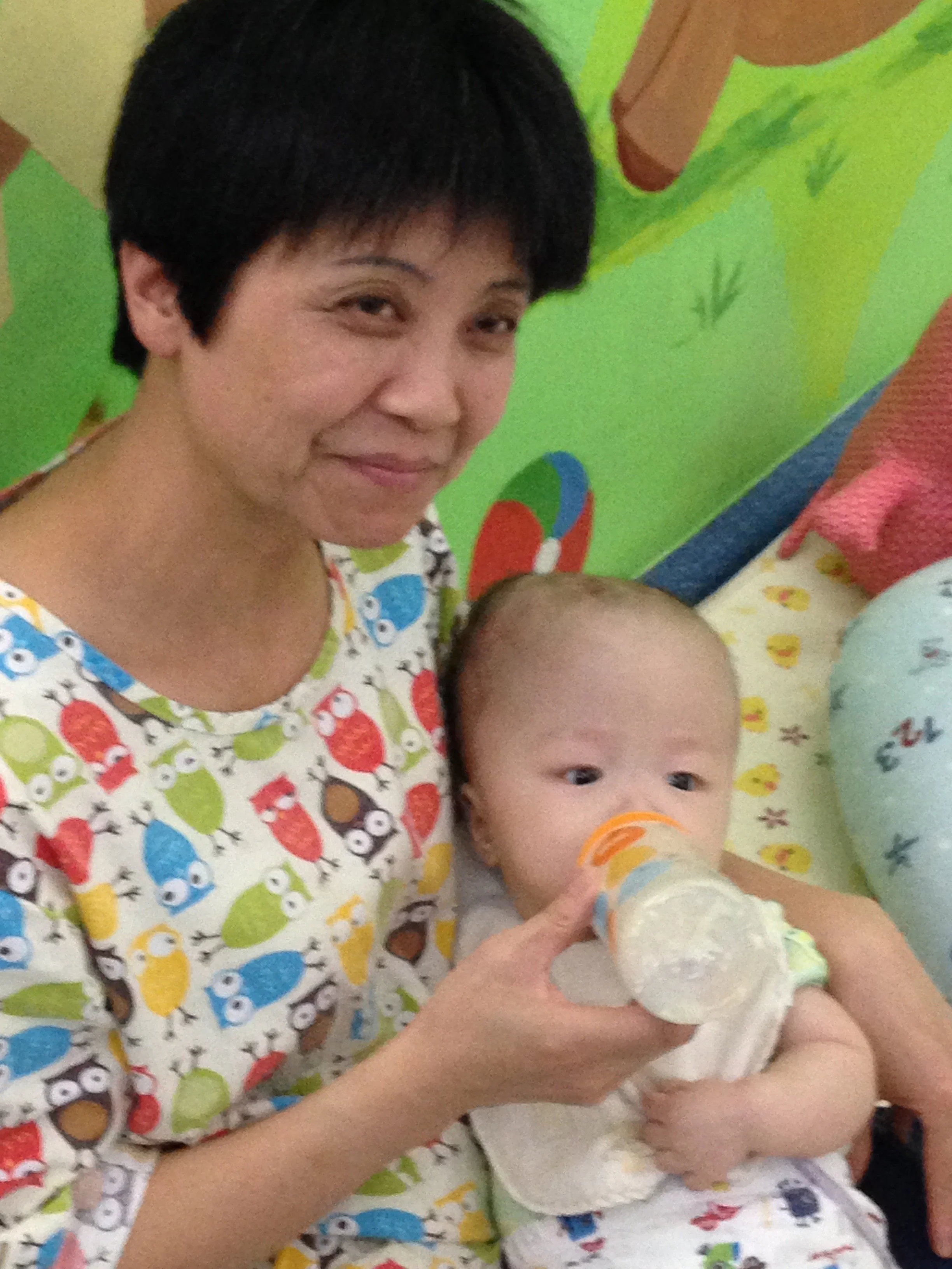 A woman with short black hair smiling, holding a baby who is drinking from a bottle. They are sitting against a colorful background with various patterns and animal illustrations.