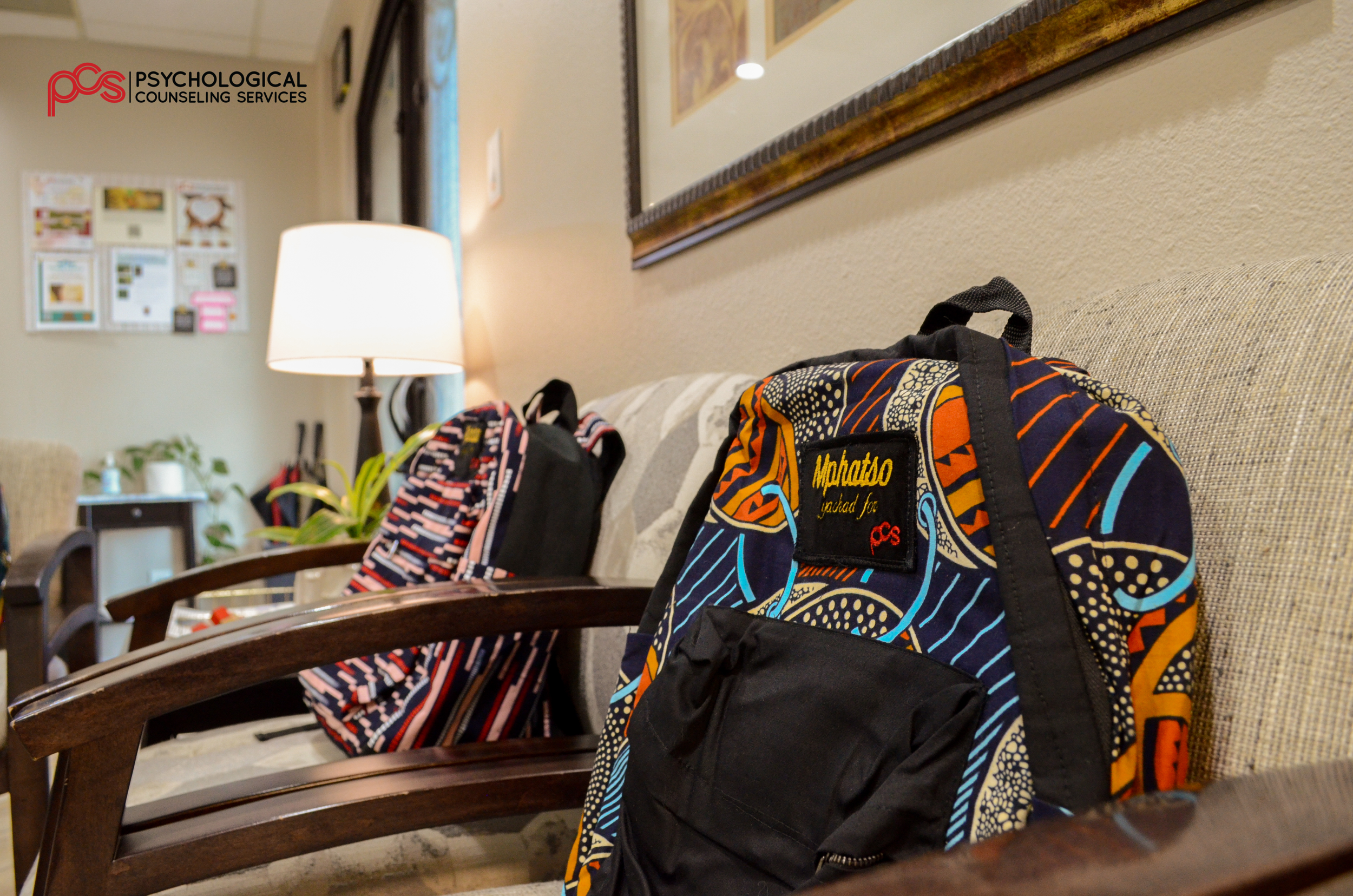 A waiting room with two colorful patterned backpacks placed on a beige cushioned bench. The room has warm lighting, a wooden chair in the foreground, a lamp, a small table with a plant and sanitizer, and framed artwork on the walls. The logo of Psychological Counseling Services is visible in the top left corner.
