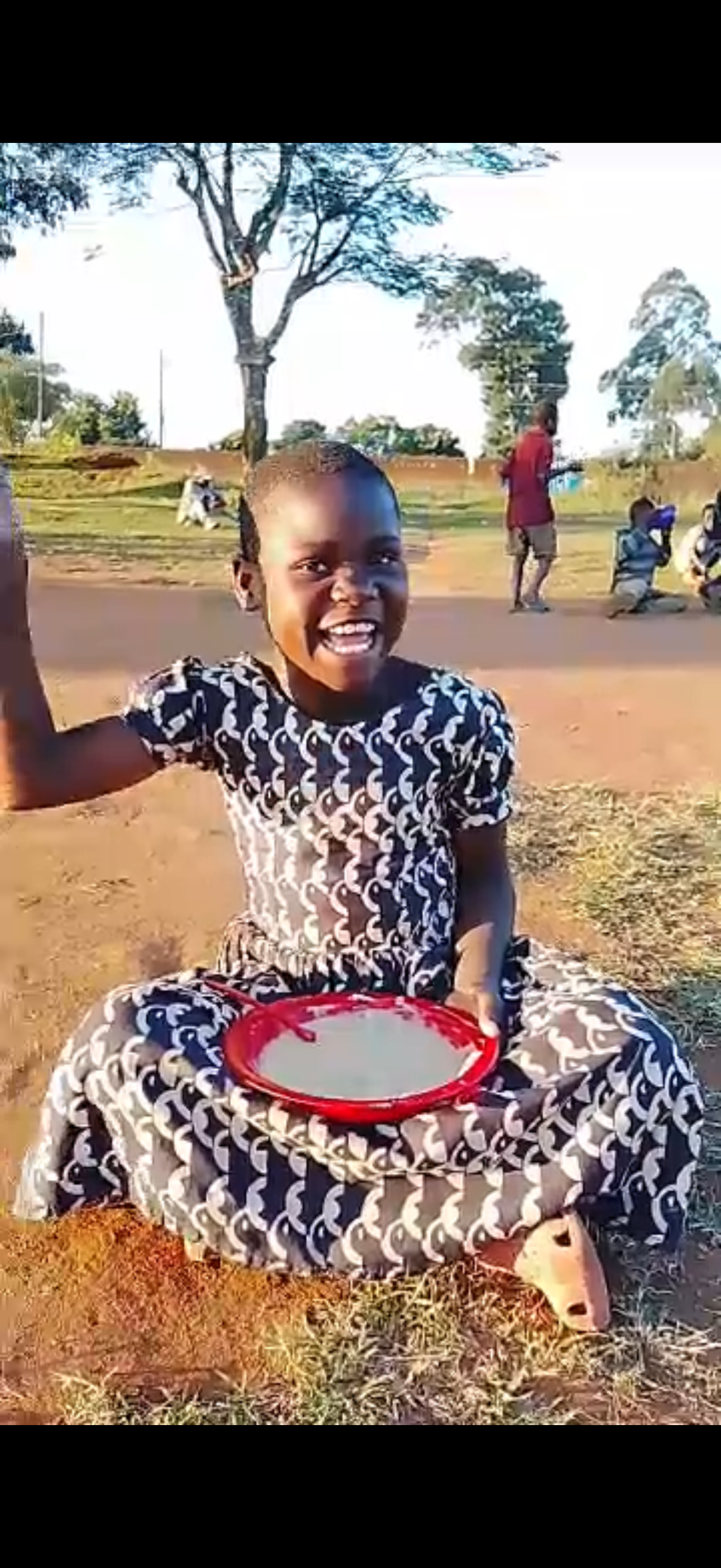 A smiling young girl wearing a black and white patterned dress, sitting on the ground with a red plate in her lap, holding a spoon, outdoors in a rural setting with trees and other children in the background.