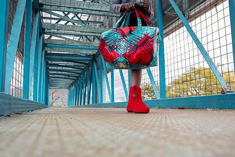 A person stands on a blue metal and wire bridge, holding a colorful bag and wearing a red sock. The photo is taken from a low angle, focusing on the sock and bag.