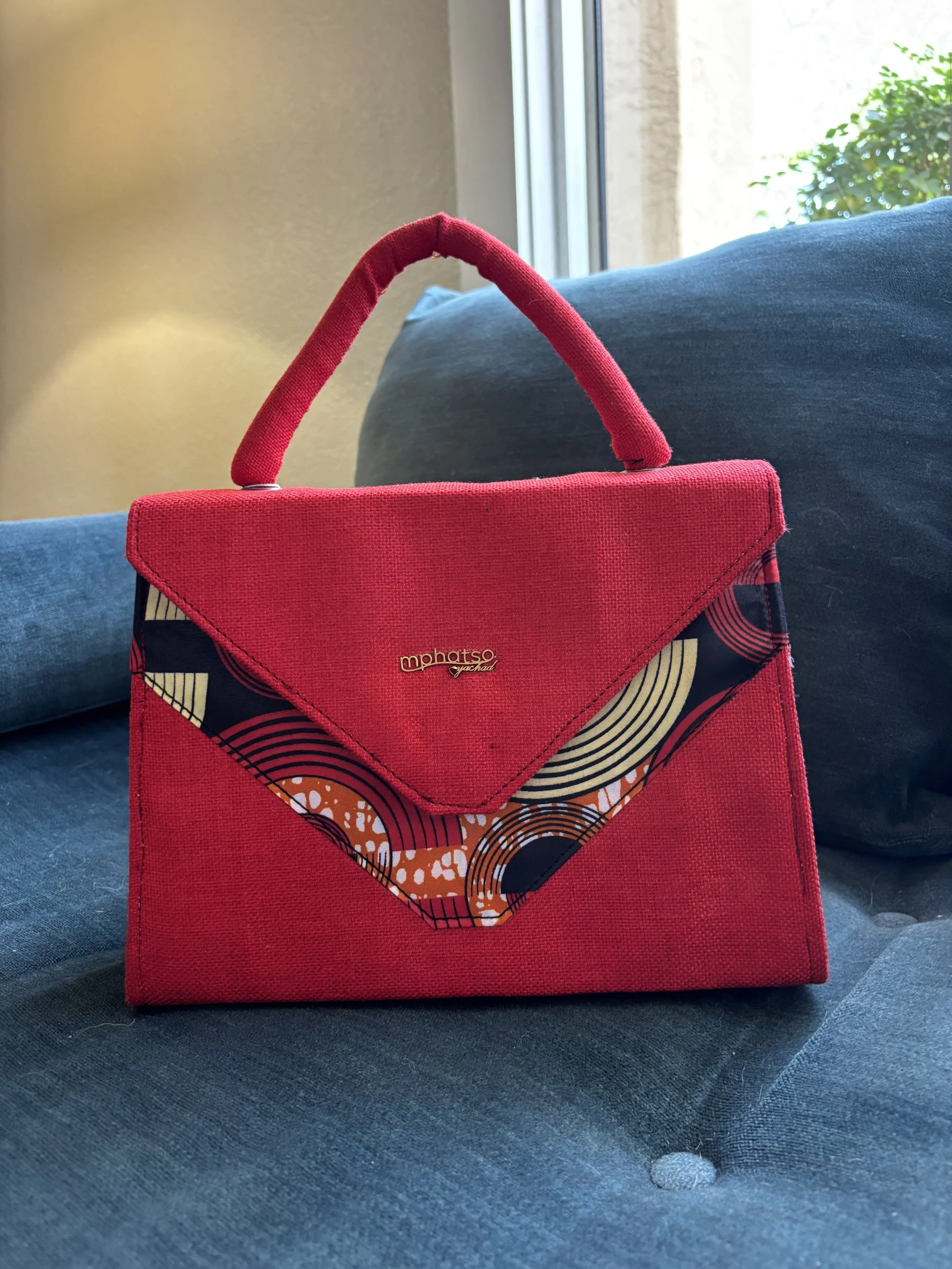 A red handbag with an abstract pattern on the front, resting on a dark blue fabric surface near a window with a green plant outside.