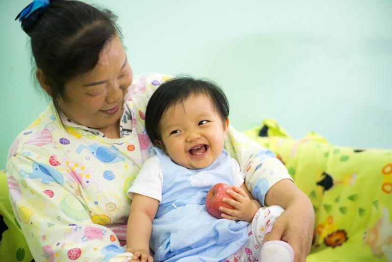 A joyful young child holding an apple while sitting on an adult woman's lap, both smiling and looking at each other, in a colorful room.