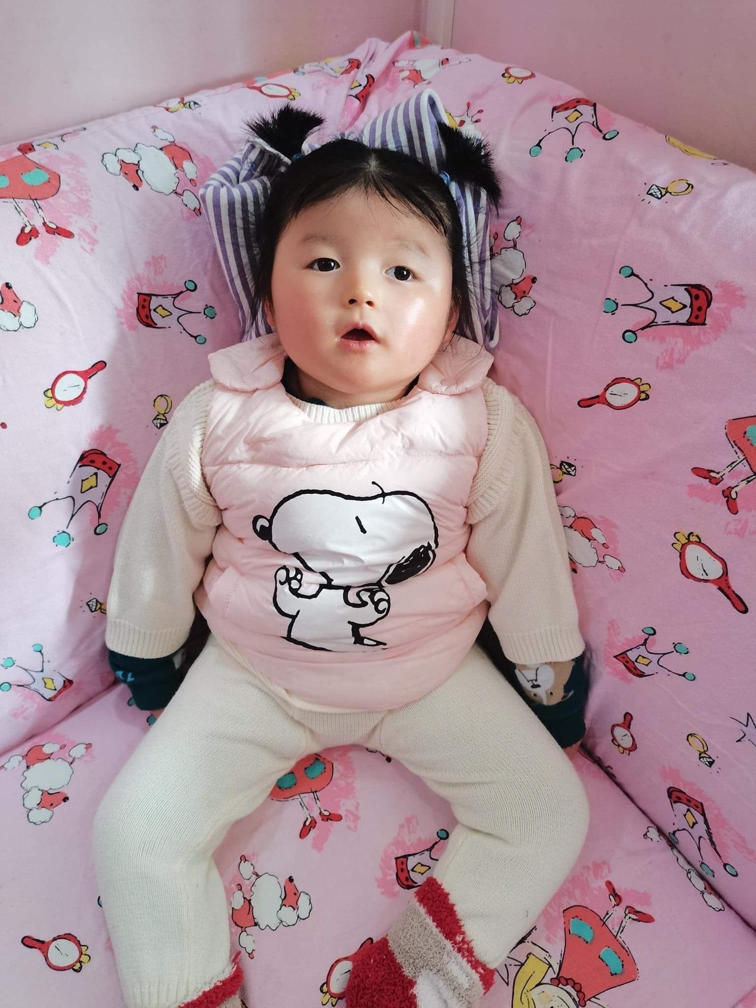 A young child with black hair and wide eyes lying on a bed with a pink sheet decorated with cartoon sheep and crowns. The child is wearing a beige sweater, cream pants, and red and beige socks with a red stripe.