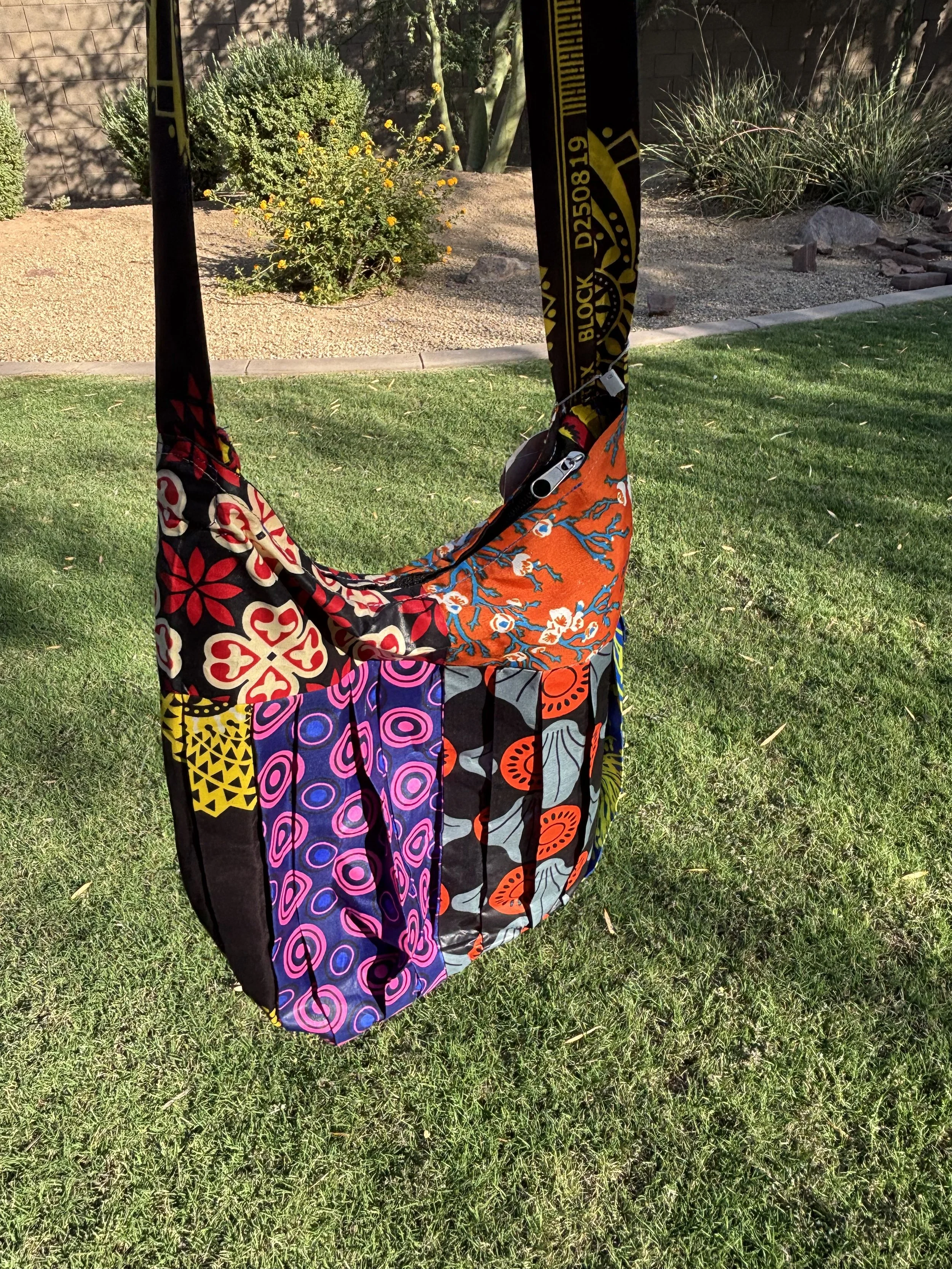 Colorful fabric sail with various vibrant patterns in orange, red, purple, black, and yellow, set outdoors on a grassy lawn with bushes and plants in the background.