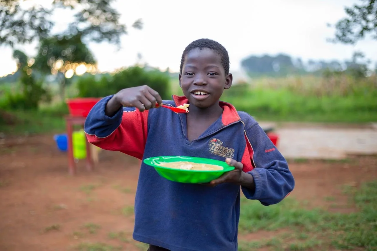 A young boy wearing a navy and red jacket is outdoors holding a green plate of food in one hand and a spoon with food in the other, smiling at the camera. The background features trees and a dirt ground.