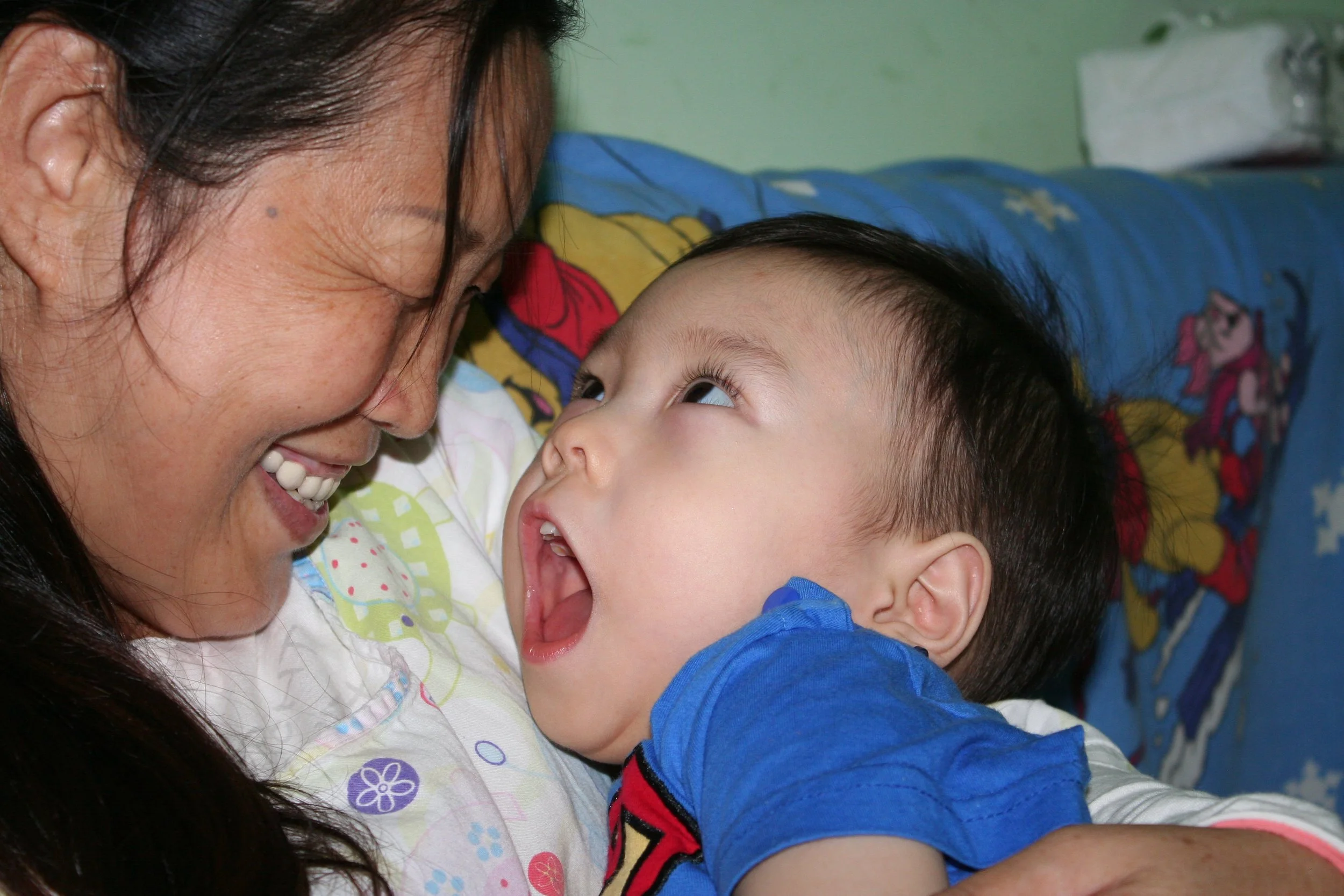 A woman and a young child lying on a bed face-to-face, smiling and looking into each other's eyes. The woman has dark hair and is wearing a white shirt with colorful patterns. The child, wearing a blue shirt with a red and black design, has short black hair. The background shows a green wall and a colorful blanket with cartoon characters.