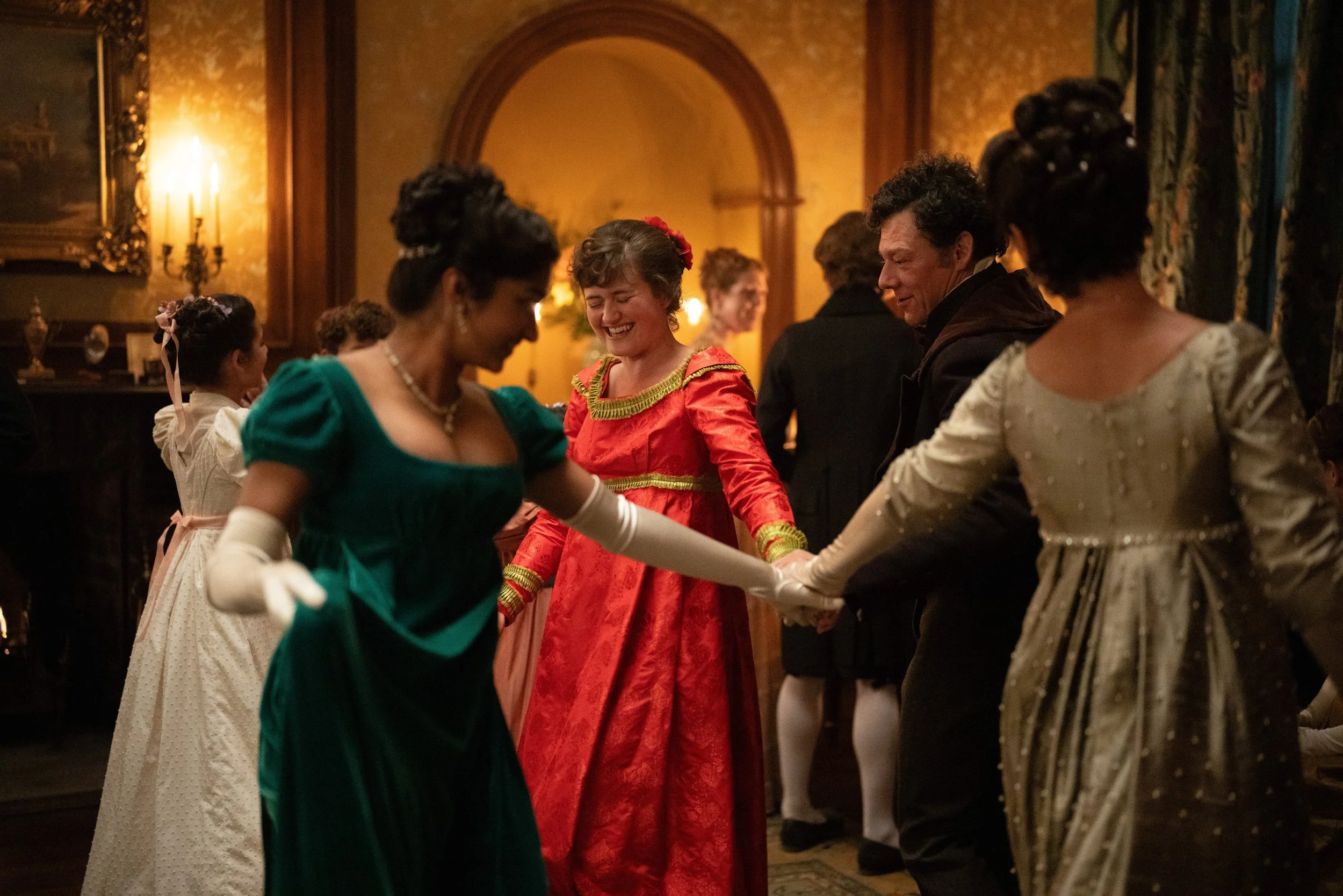 People dressed in historical costumes dancing in a semi-circle, holding hands in a Victorian-style room with ornate woodwork and warm lighting.