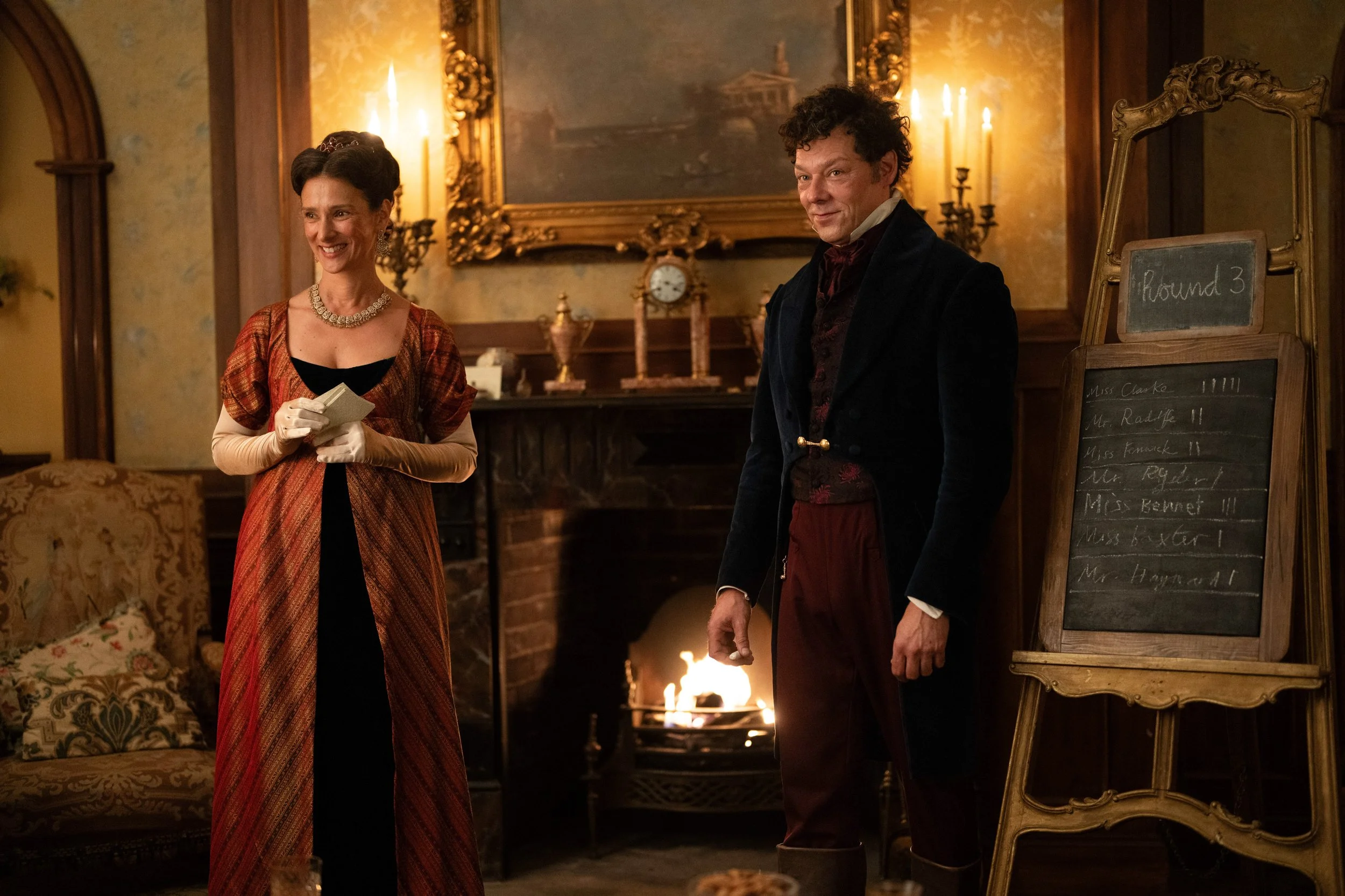 Two people dressed in period clothing, a woman holding a notepad and a man standing beside a chalkboard, in a room with a fireplace and ornate decorations, resembling a historical setting.