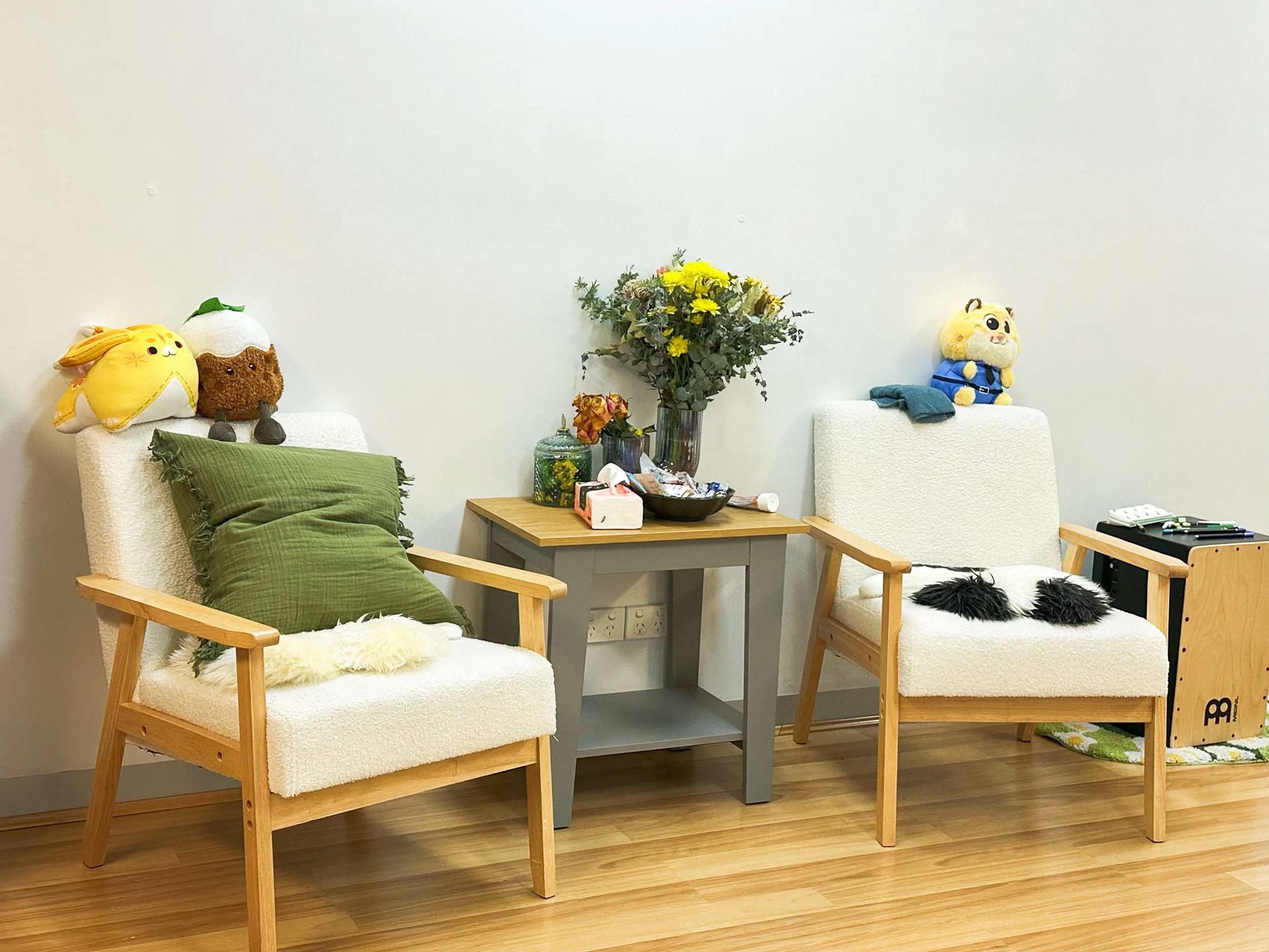 Cozy living room with two white armchairs, one with green cushions, and plush toys on the backrest, a small wooden side table with flowers, snacks, and a tissue box, and a plain white wall background.