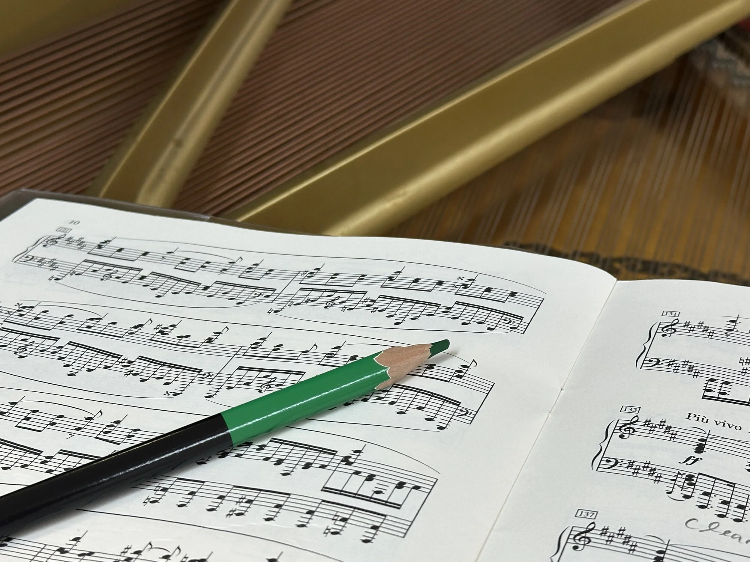 Sheet music with musical notes, a green and black pen, and a pencil resting on the music paper, with part of a piano visible in the background.