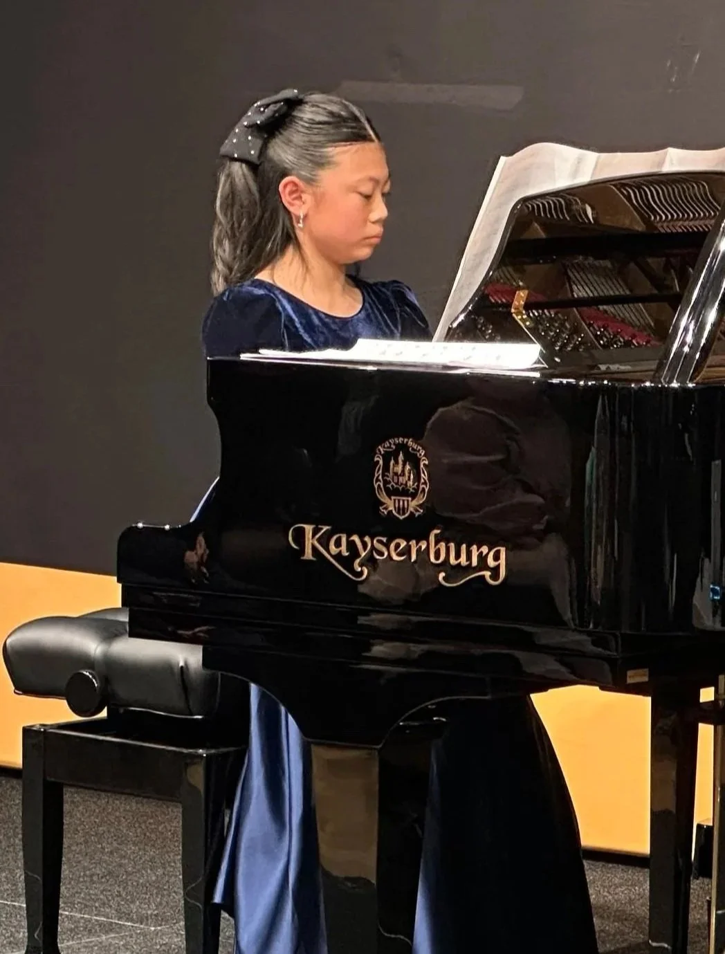 A young girl playing a black Kayserburg grand piano with sheet music on top, wearing a dark blue dress and a black bow in her hair, in a room with dark walls.