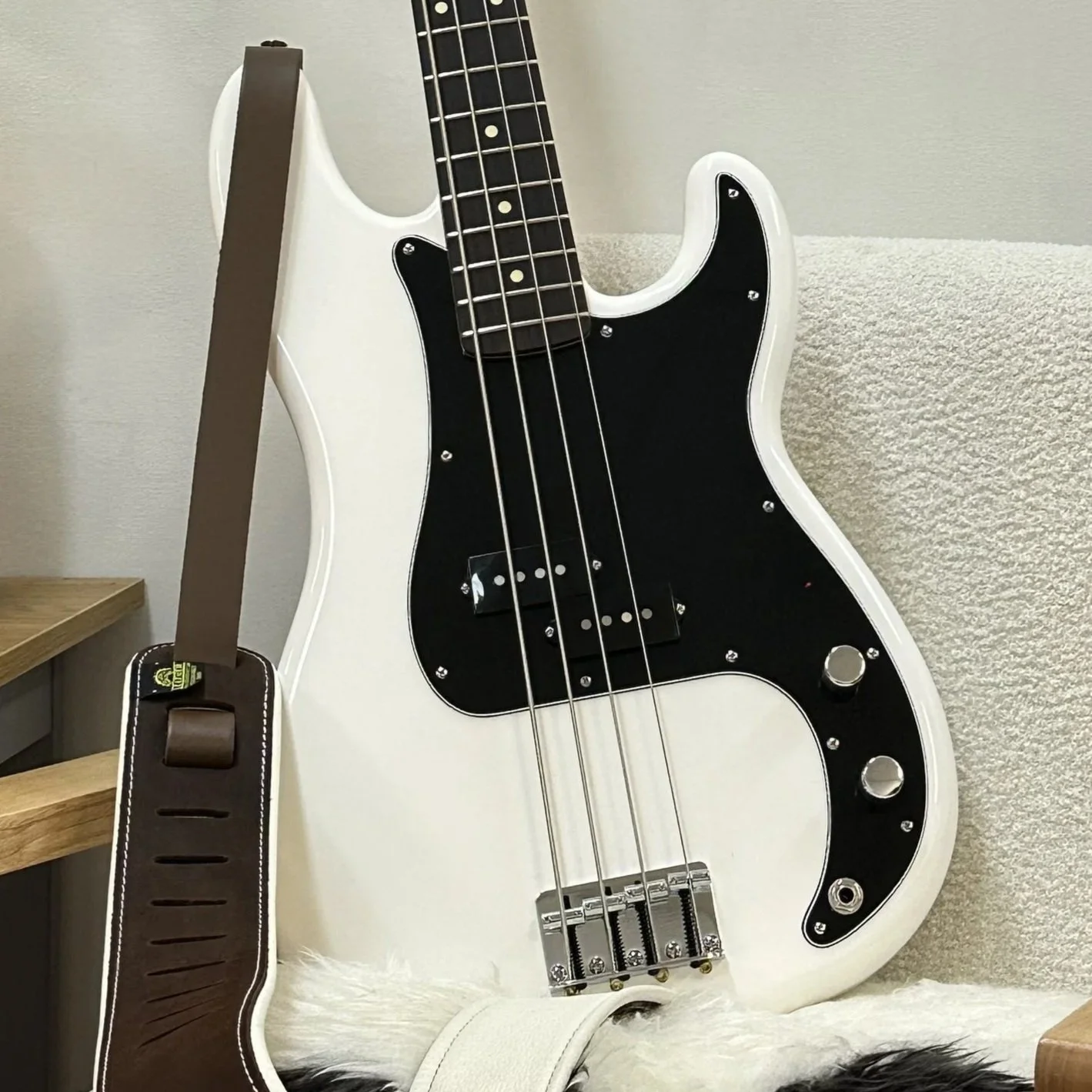 A white electric bass guitar with black pickguard resting against a light-colored wall, with a brown strap hanging over it, and a small wooden shelf to the left.