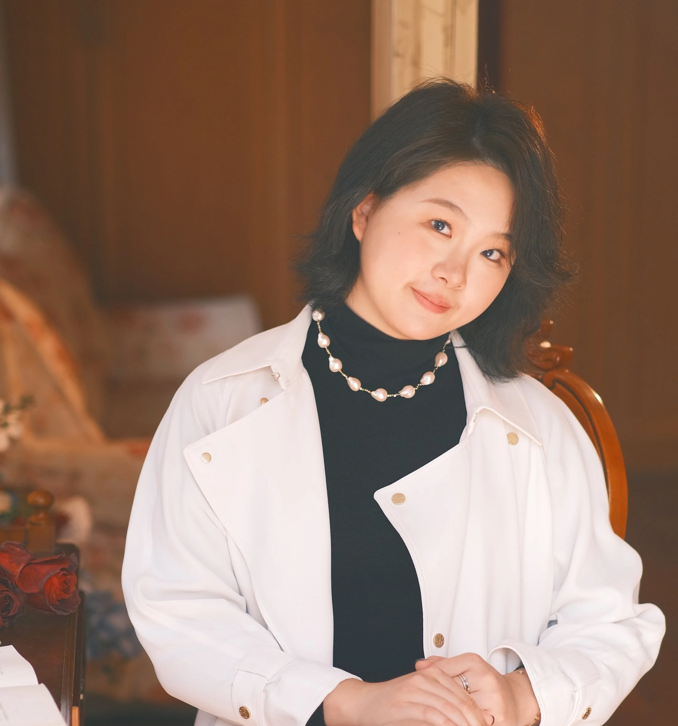 A woman with black hair and light skin sitting at a table, wearing a white blazer over a black top, with pearl jewelry, and a warm smile, in a cozy indoor setting with a wooden background.