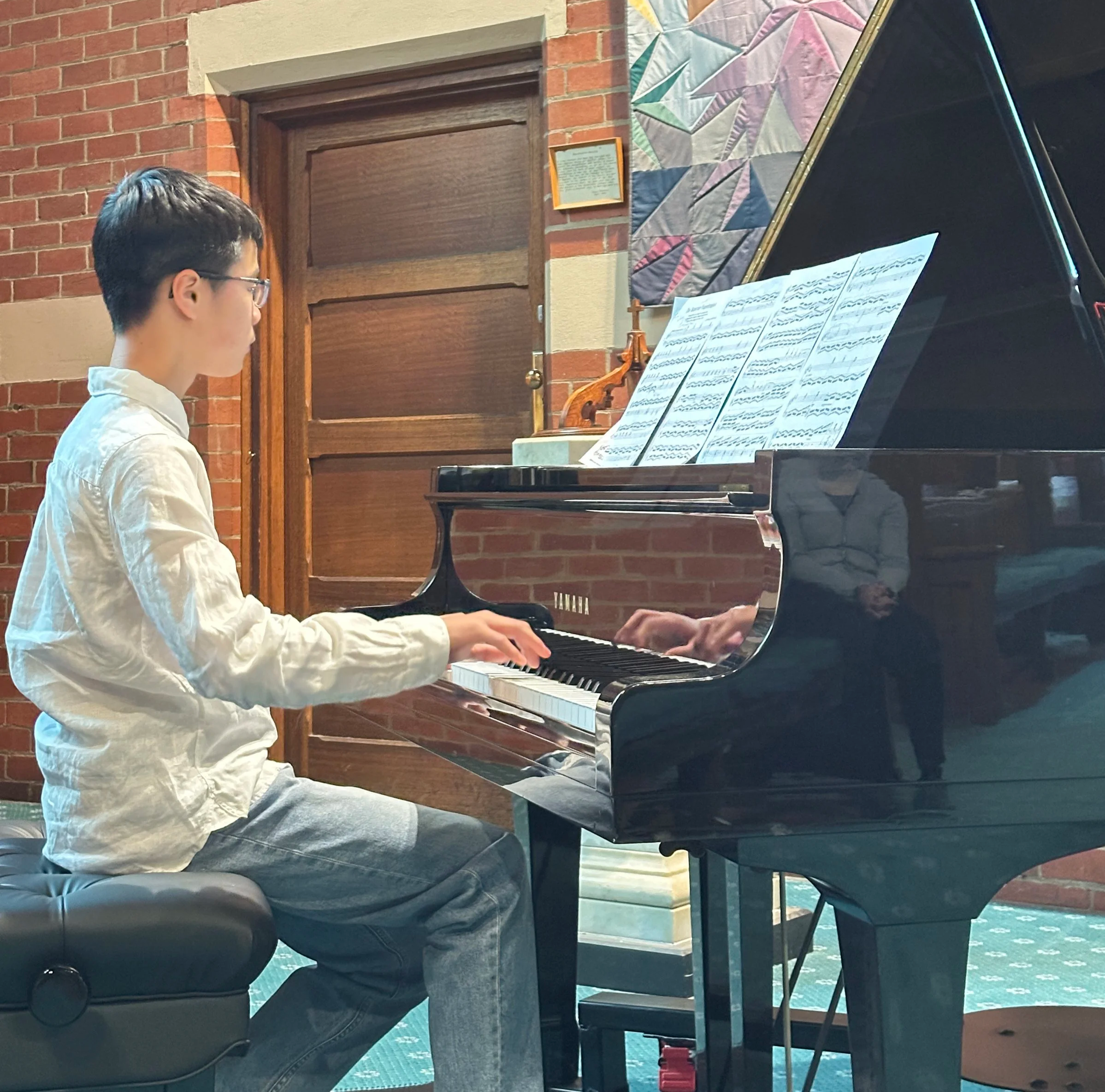 A young man with glasses playing a grand piano indoors, sitting on a black padded bench. The background features a brick wall, a wooden window frame, and colorful quilted artwork on the wall.