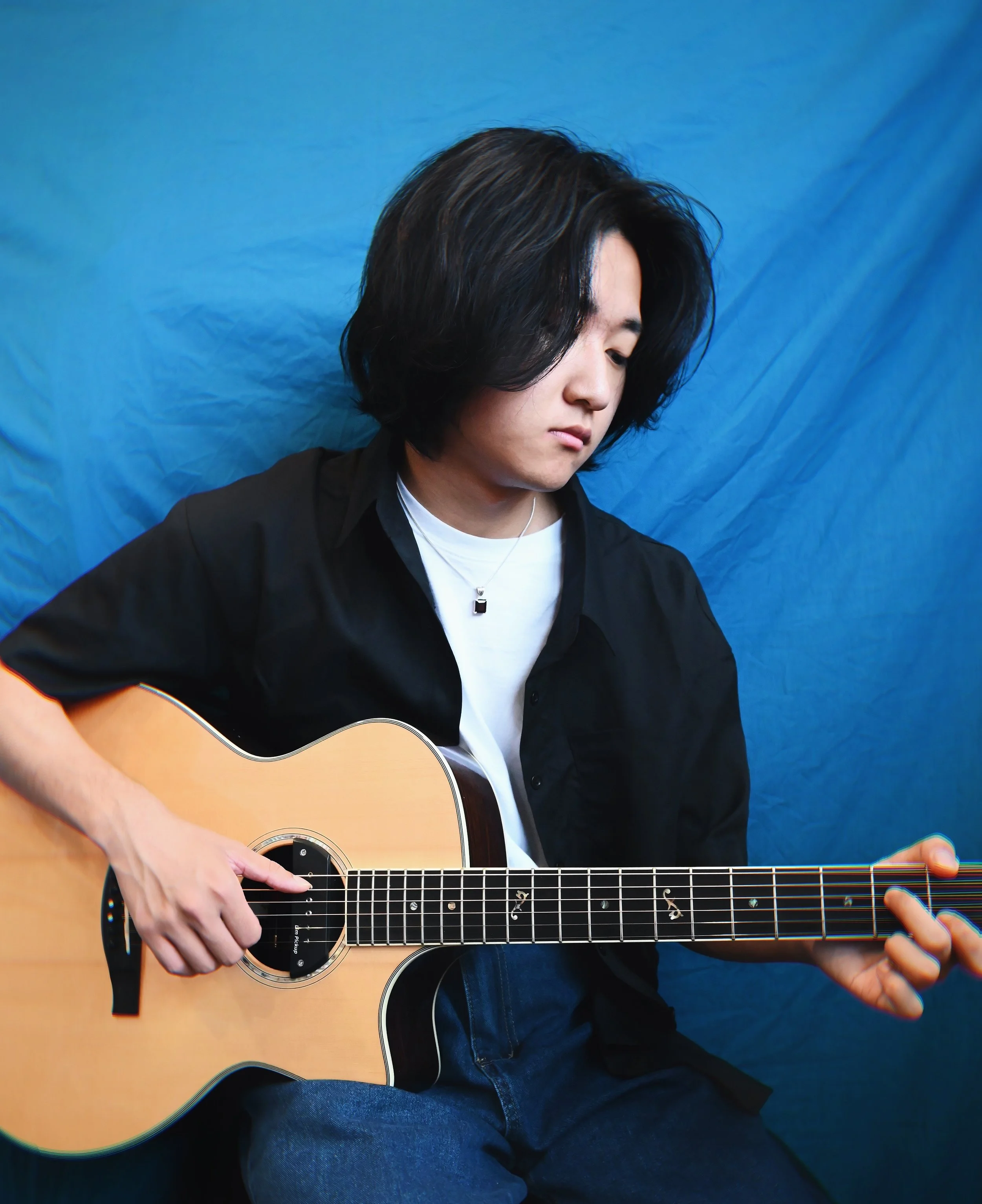 Person playing acoustic guitar with a blue background, wearing a black shirt and jeans