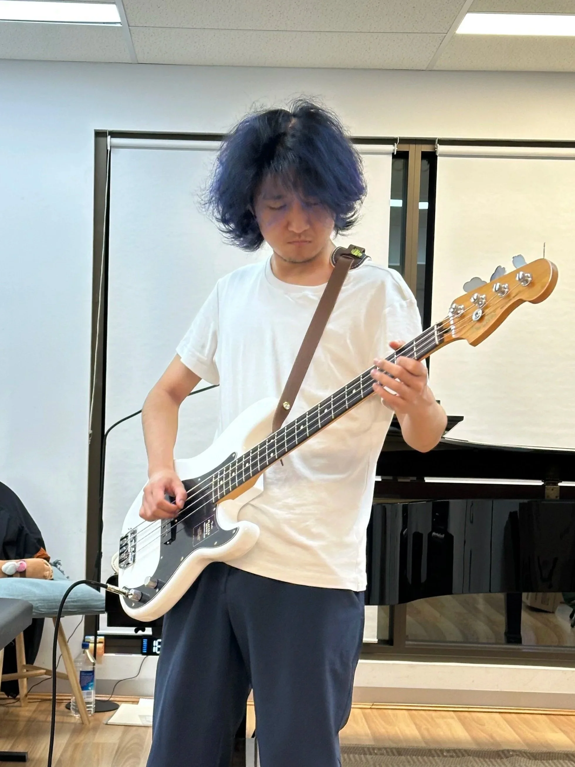 A person with curly dark hair playing a white electric guitar indoors, with a piano in the background.