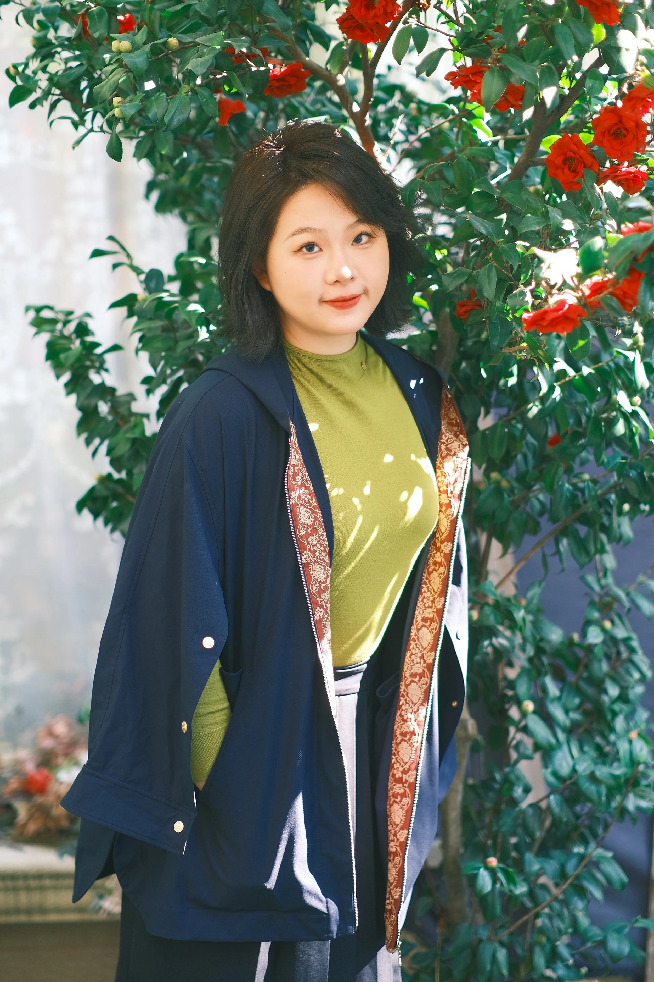 A young woman with black hair in a layered cut, wearing a green shirt and a navy blue jacket with a patterned lining, standing in front of a lush green bush with red flowers, outdoors in sunlight.