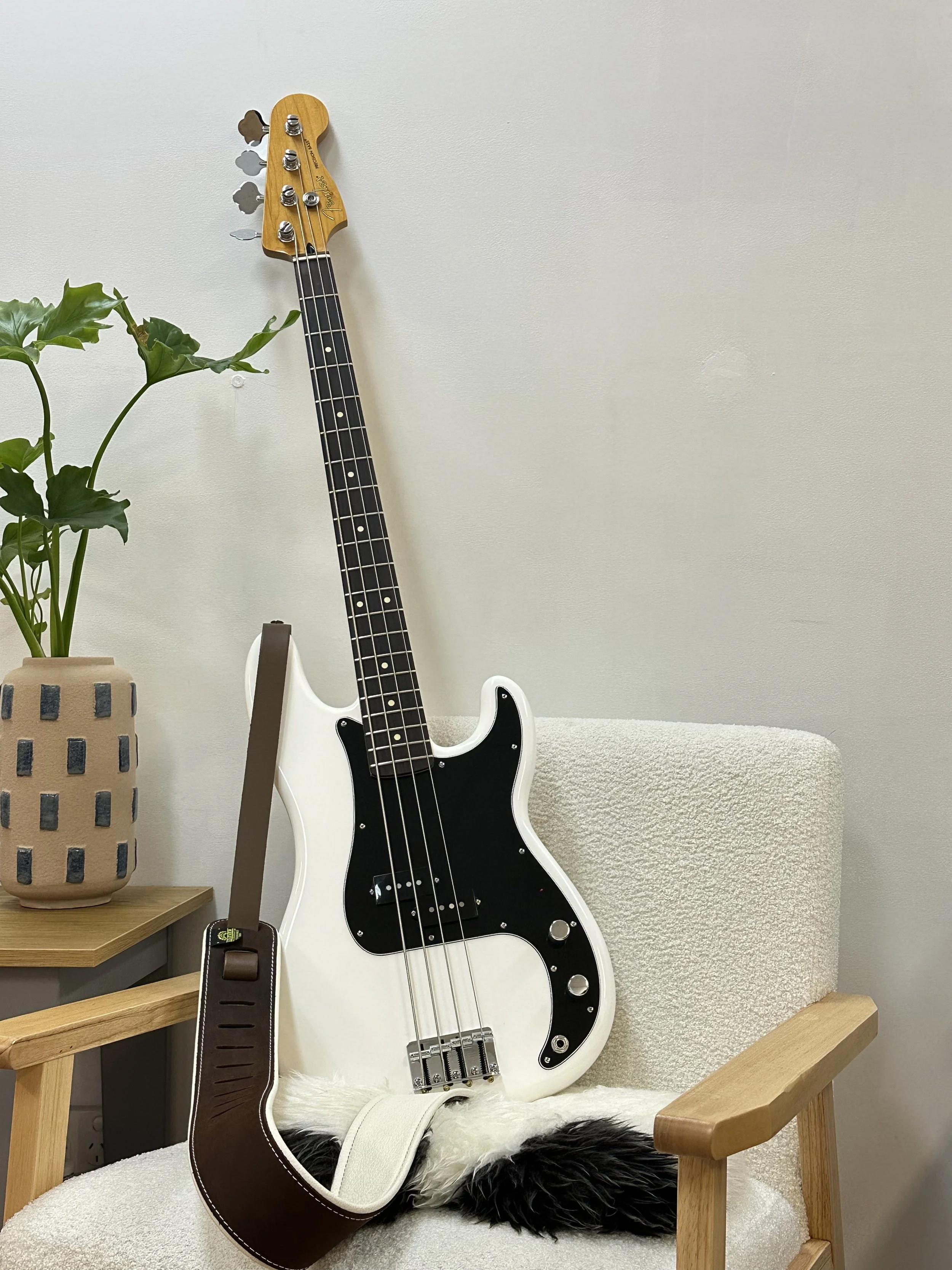 White electric bass guitar resting on a beige armchair with a black and white fuzzy pillow. A brown strap is draped over the guitar. To the left, there is a beige pot with a green leafy plant on a wooden side table.