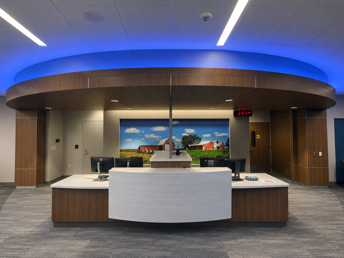 Modern reception desk in an office with computer monitors, a large digital clock showing 6:54, and a farm landscape painting in the background.