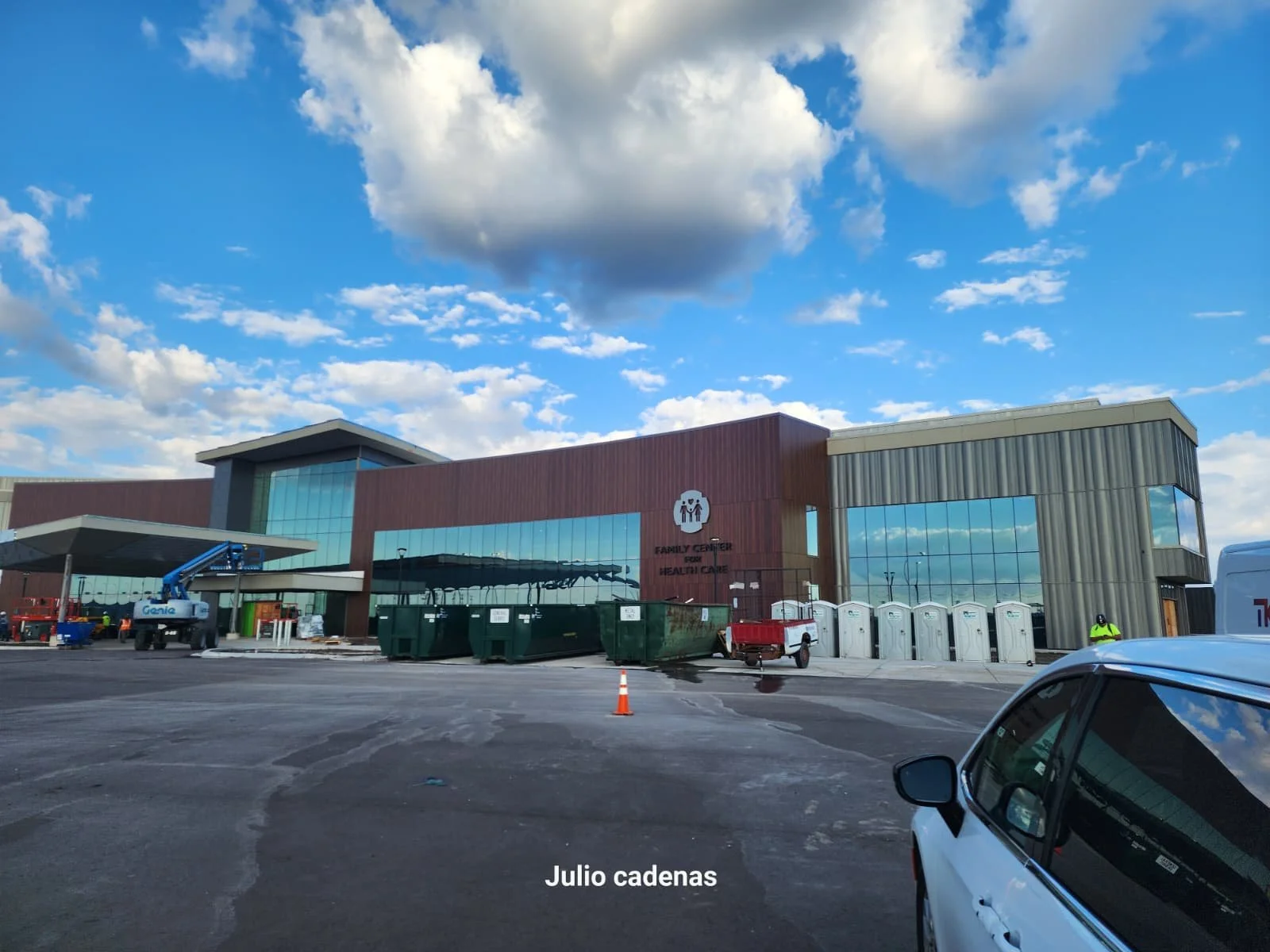 The exterior of a modern family health center with construction or maintenance work active, including a crane, construction materials, and portable toilets, with a bright sky and clouds overhead.