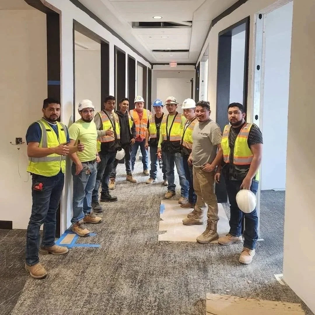 Group of construction workers and contractors standing in a hallway of a building under construction or renovation, with some holding helmets and wearing safety vests.