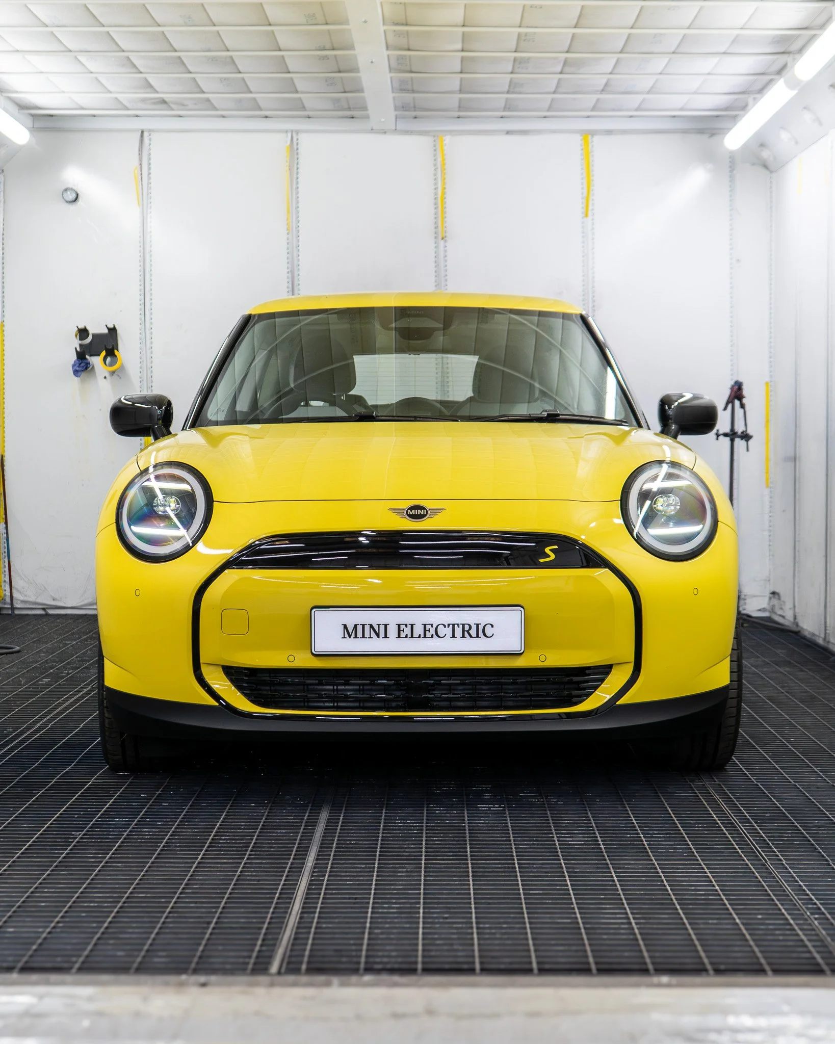 Yellow Mini Electric car inside a clean, white, well-lit garage or workshop with tools hanging on the wall, and a sign on the front bumper reading 'MINI ELECTRIC'.
