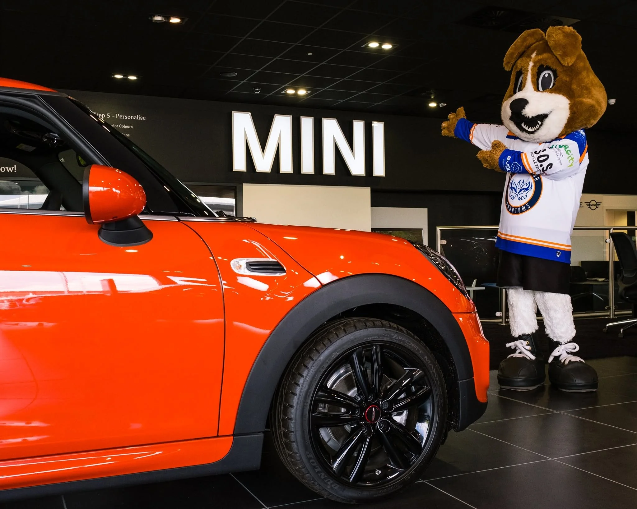A person in a dog mascot costume wearing a hockey jersey standing next to a red mini car inside a showroom with a large illuminated 'MINI' sign on the wall.