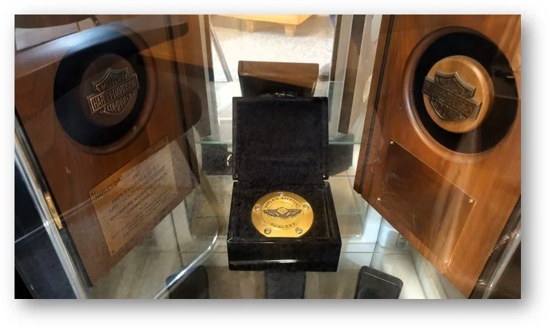 A Harley-Davidson commemorative plaque displayed on a black podium, flanked by two wooden Harley-Davidson logos mounted on plaques.